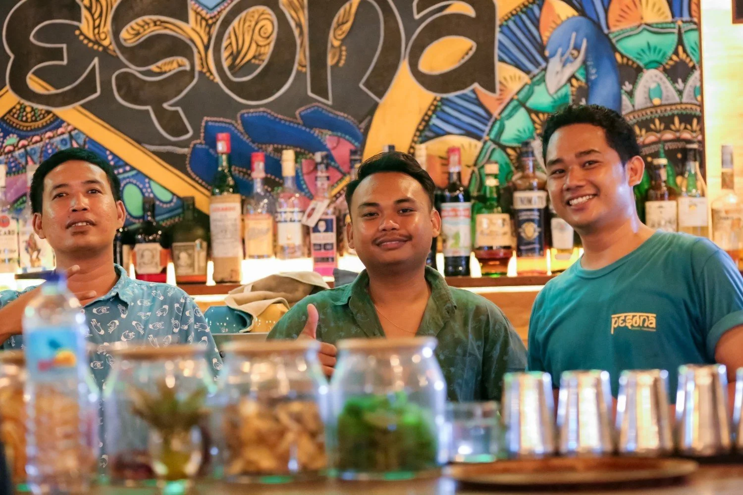 Three men sitting at a bar with bottles of liquor behind them, smiling and posing for the camera, with colorful mural artwork on the wall.