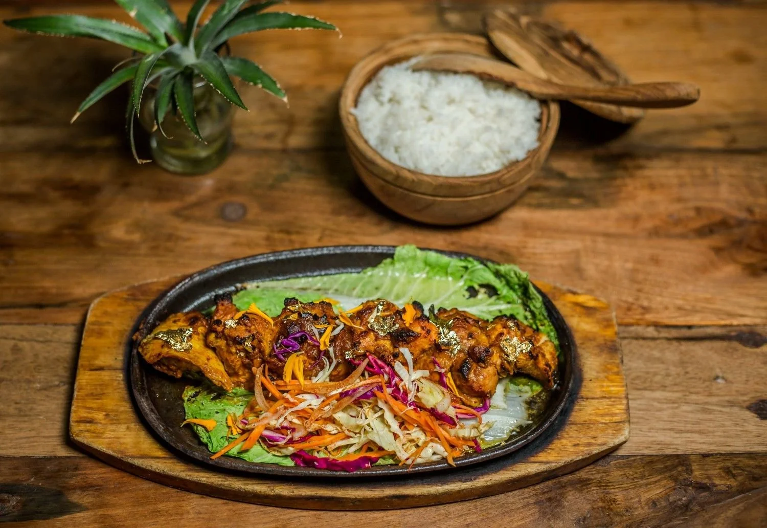 A plate of grilled tandoori-style chicken served with fresh salad on a wooden board, with a bowl of rice in the background.