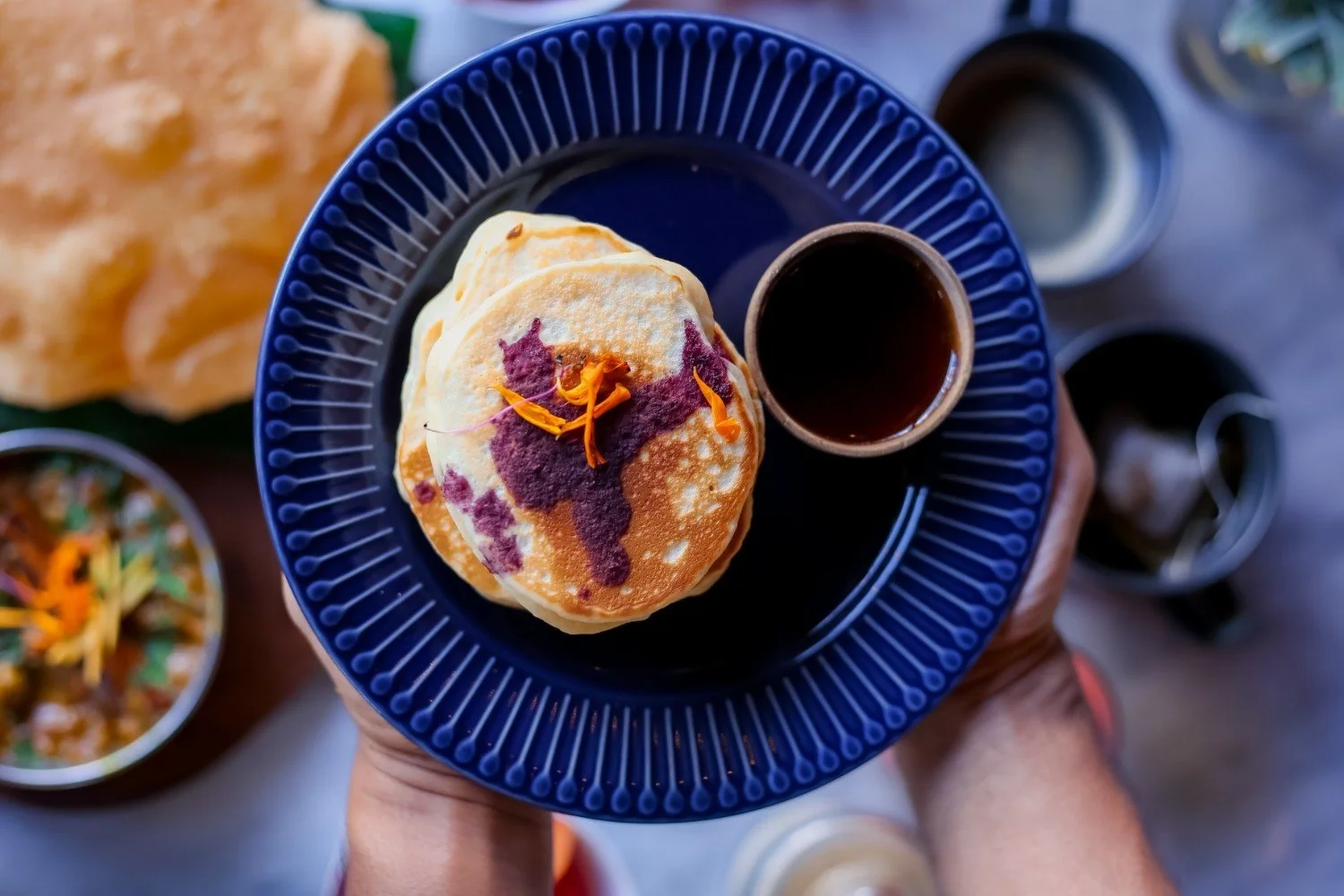 Stack of fluffy pancakes topped with purple ube sauce and edible flower petals, served on a blue plate with a cup of coffee.