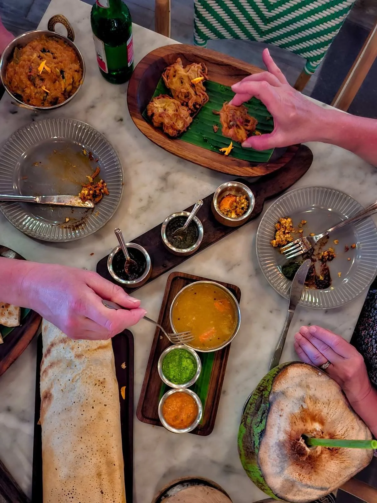 Overhead view of an Indian meal on a marble table with dosa, curry, pakoras, chutneys, coconut water, and several people sharing food.