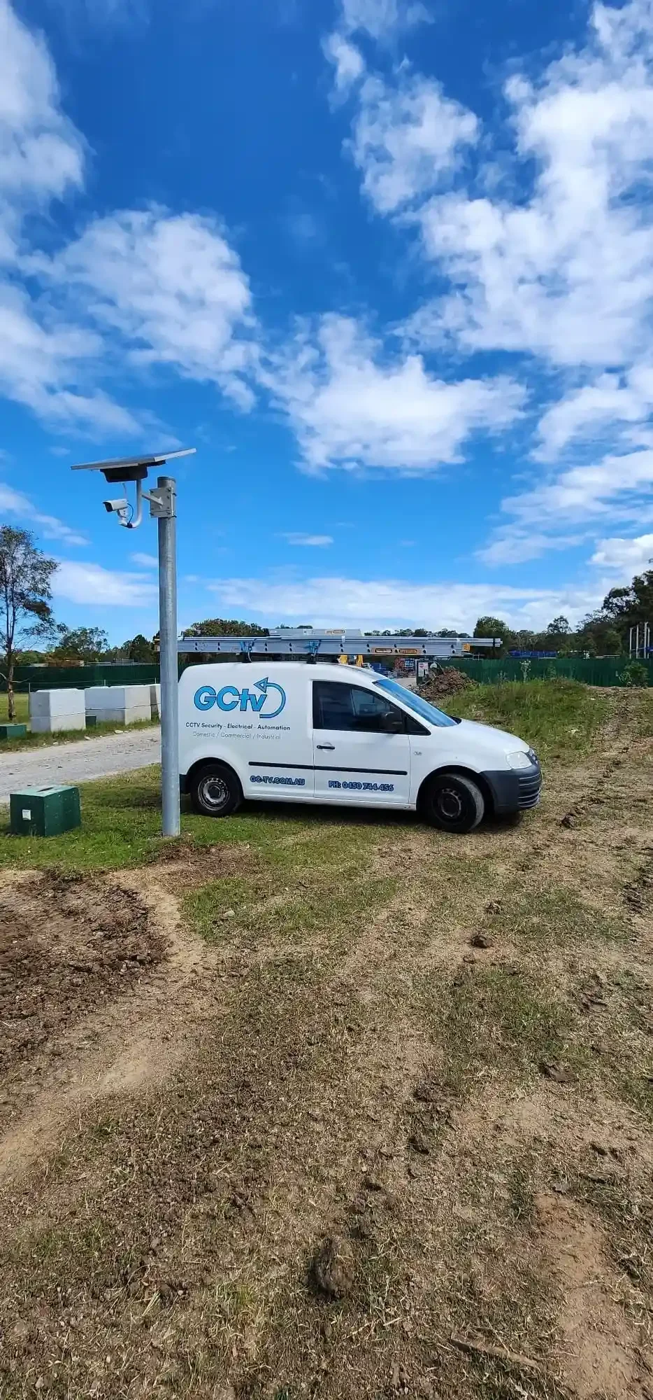 A white GCTV service van parked on a grassy and dirt area near a CCTV pole. The sky is blue with scattered clouds, and there is construction or development activity in the background.
