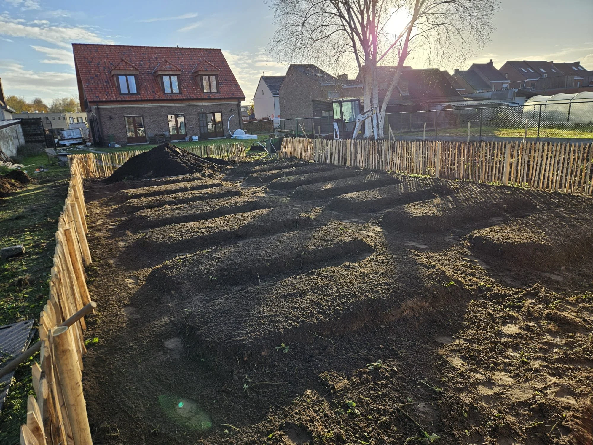 Een moestuin wordt voorbereid met uitgegraven grond en een houten omheining, er staat een graafmachine en er is een huis op de achtergrond, onder een heldere blauwe hemel met de zon die schijnt.