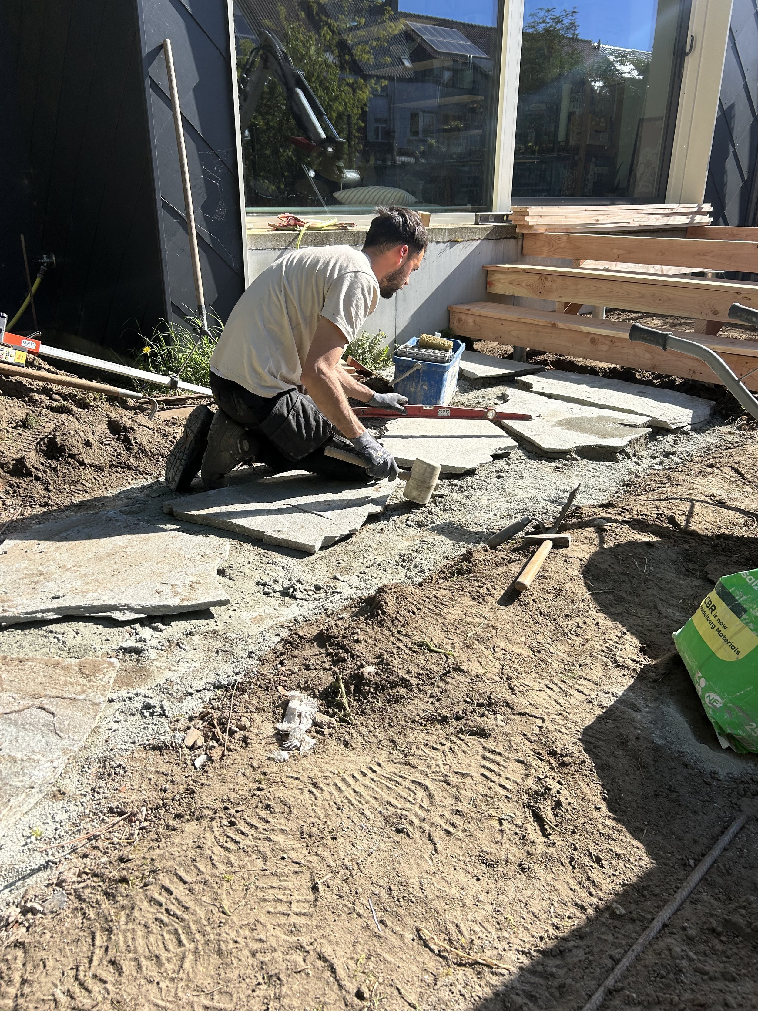 Man legt grote flagstones neer voor een trap bij een huis onder een blauwe lucht.