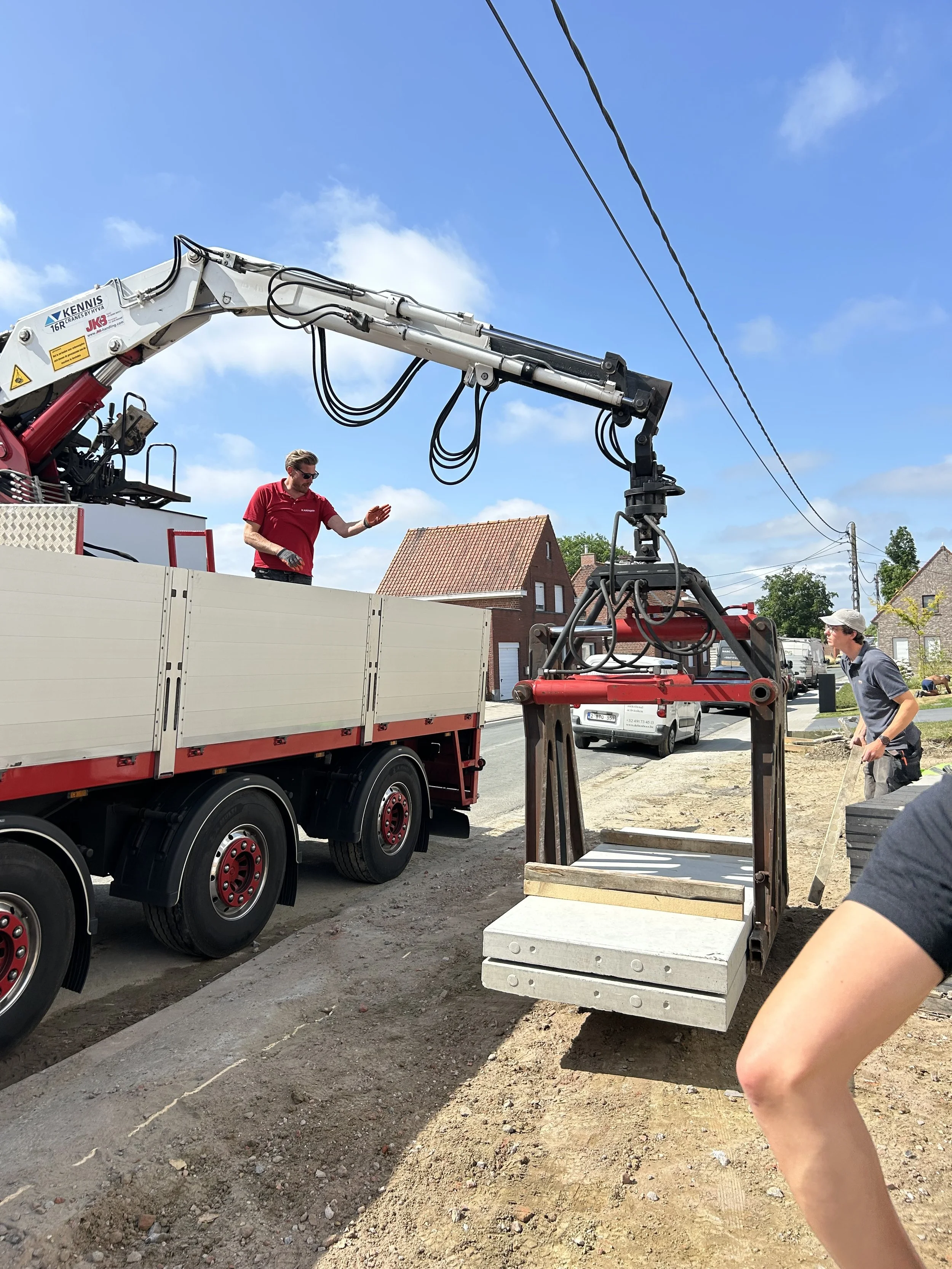 Twee mannen werken op een bouwplaats met een grote kraan die een betontegel optilt, terwijl een andere man op de achtergrond staat en meewerkt. Er zijn huizen en een blauwe hemel te zien.