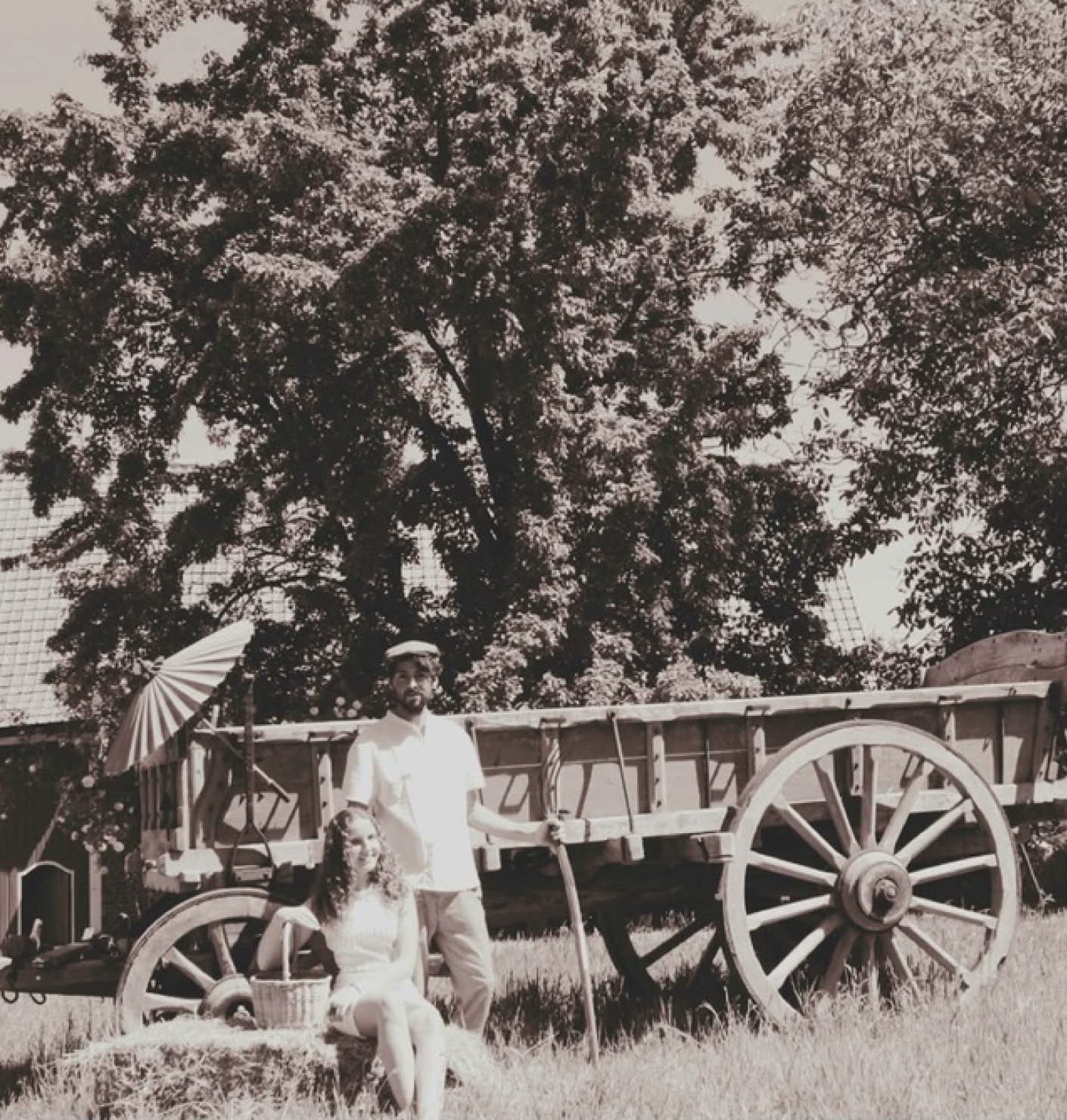 Een man en een vrouw staan voor een houten wagen met grote wielen onder een grote boom, de man houdt een stok vast, de vrouw zit op het gras, er hangt een  parasol aan de wagen.