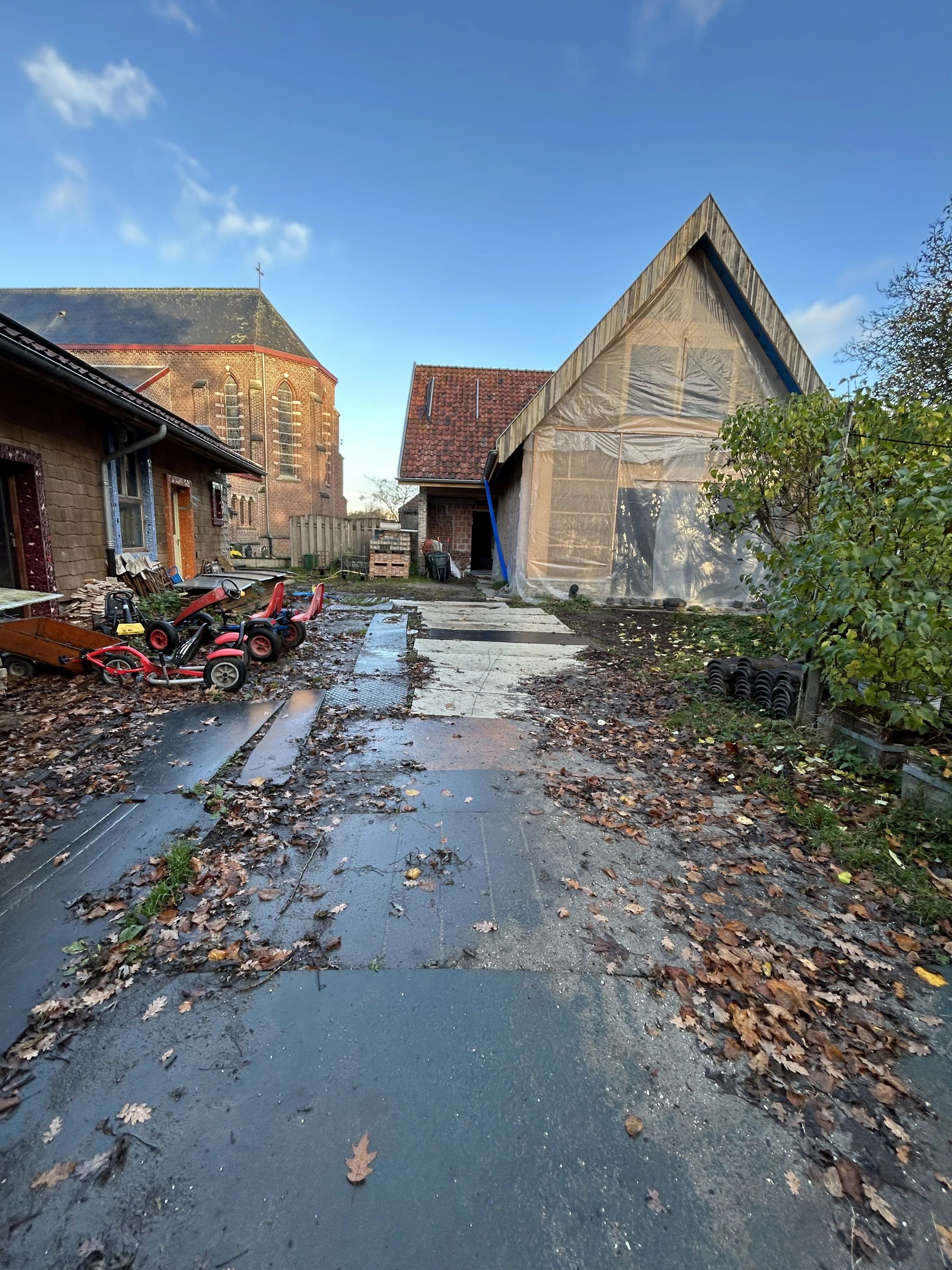 De foto toont een werf van  twee houten huizen. Een met plastic bedekking en een stenen huis met een rode dak. In de tuin liggen speelgoedauto's en andere tuingereedschappen.