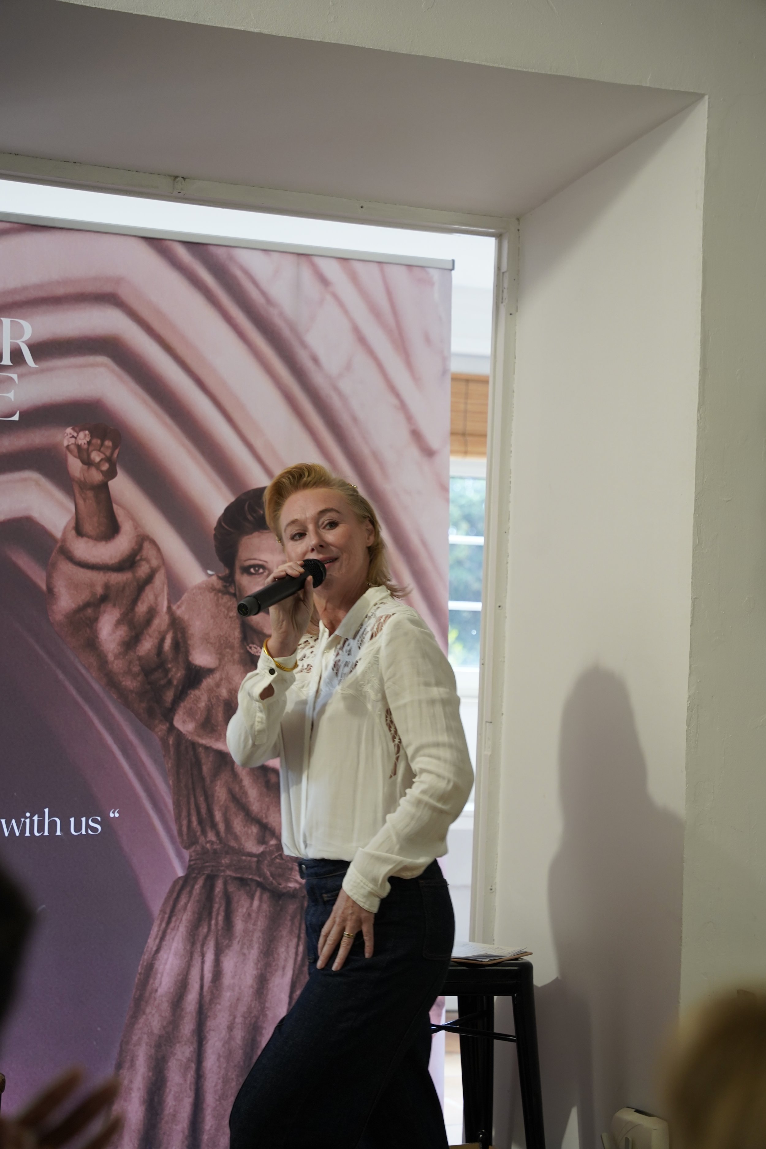 A woman with blonde hair, wearing a white blouse, stands in front of a poster, speaking into a microphone, in a room with white walls and a window in the background.