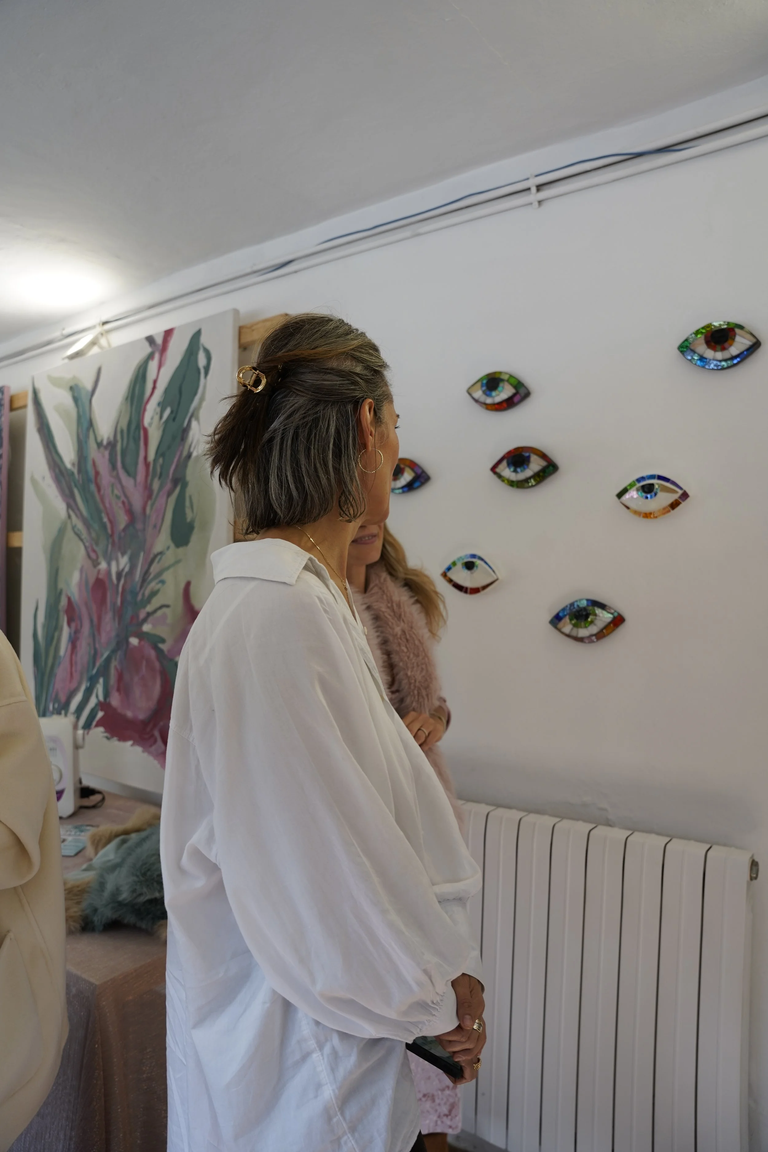 A woman with dark, shoulder-length hair, wearing a white shirt, standing in an art gallery or studio, looking at wall-mounted decorative eyes made of colorful mosaic tiles.