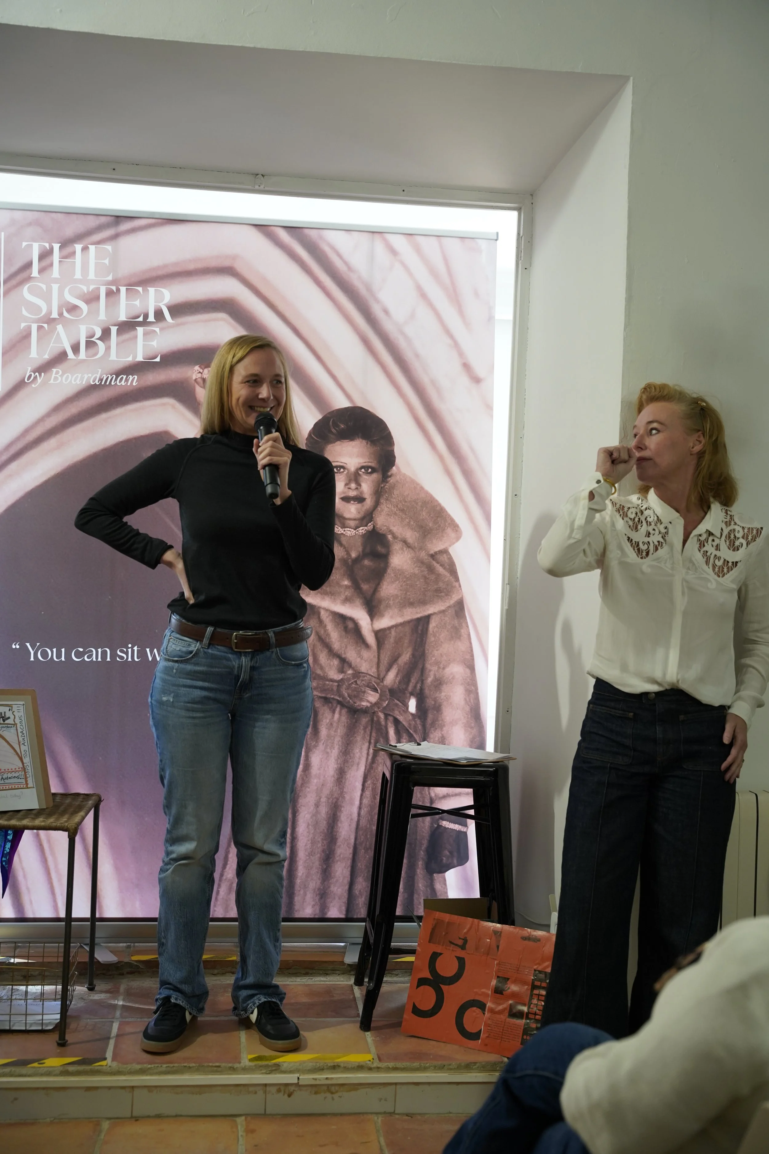Two women standing in front of a large poster with the title 'The Sister Table by Boardman.' One woman is speaking into a microphone, and the other is looking at her while touching her face. The scene appears to be at an event or presentation.