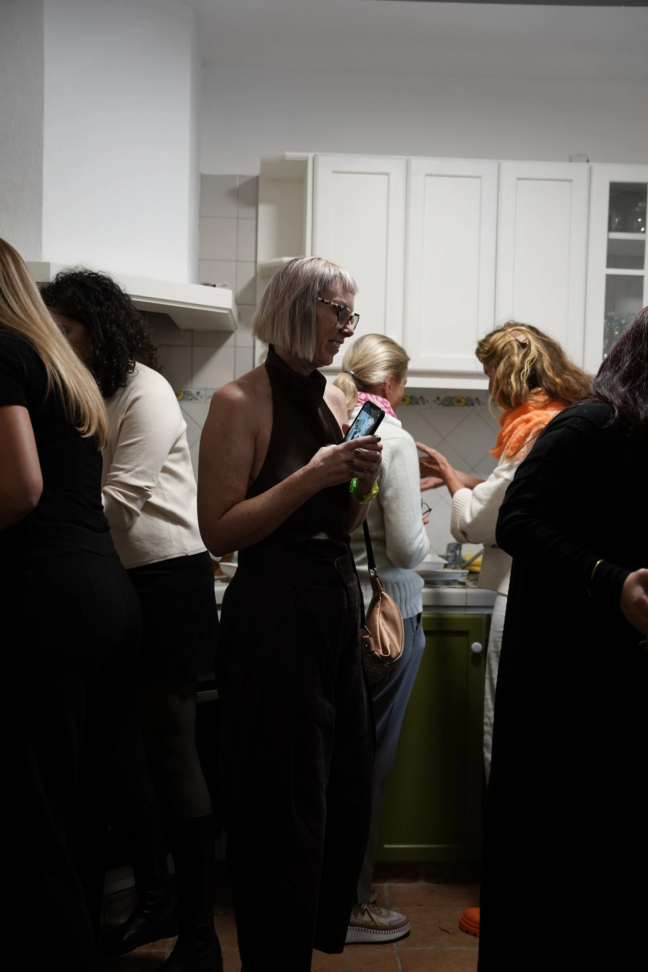 A group of women standing closely together in a kitchen, some looking at their phones, others engaged in conversation, with white cabinets and tiled backsplash in the background.