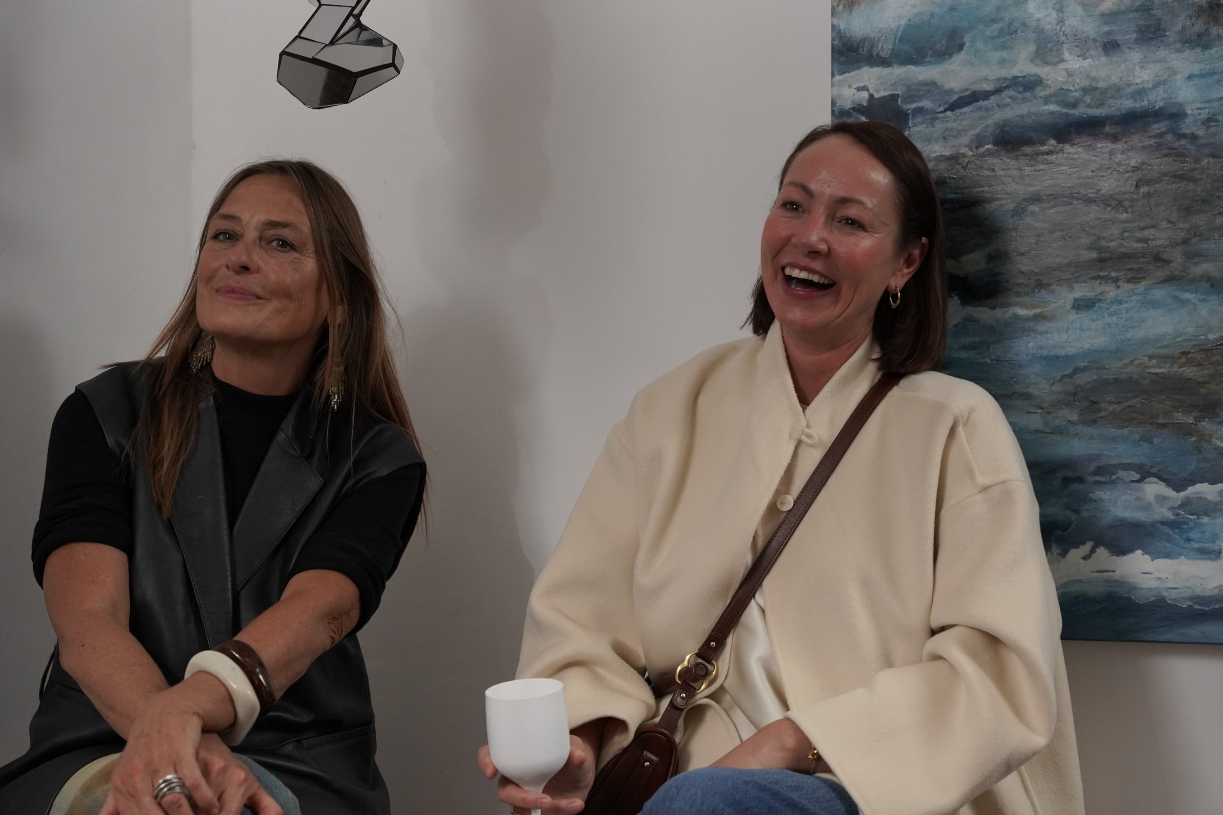 Two women sitting together, smiling and laughing. One woman has long brown hair and is wearing a black leather vest, the other has short brown hair, wearing a cream-colored coat and earrings. They are seated in an art gallery with a painting of ocean
