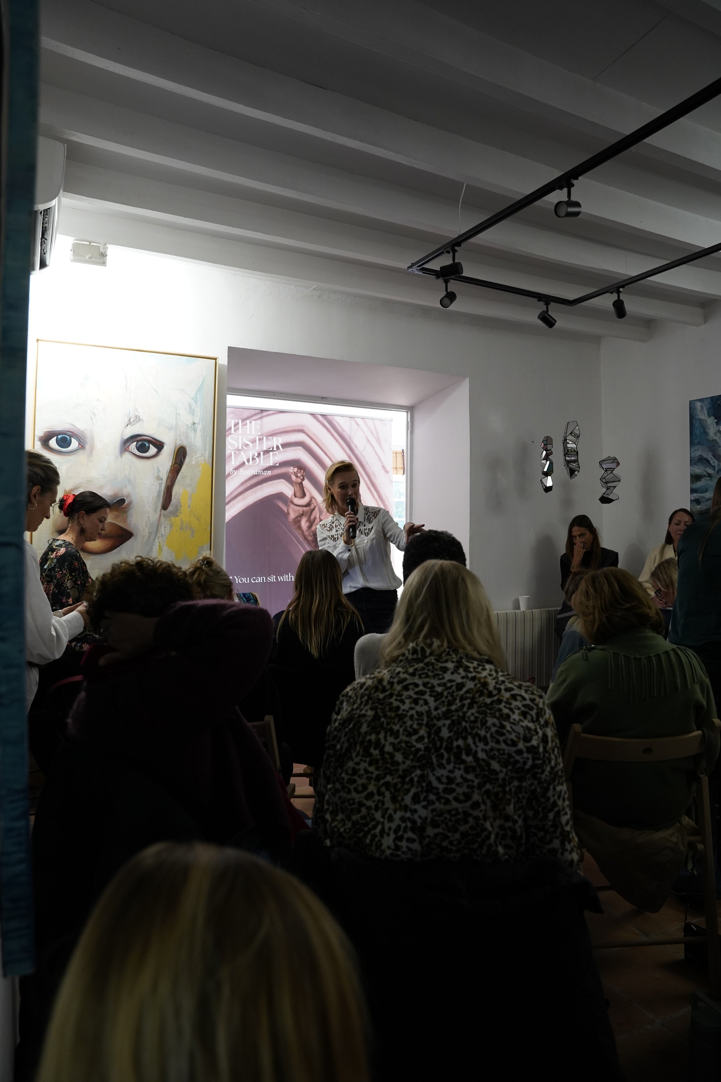A woman speaking into a microphone at a presentation event, with large artwork and hanging decor on the wall behind her, and an audience seated in front of her.