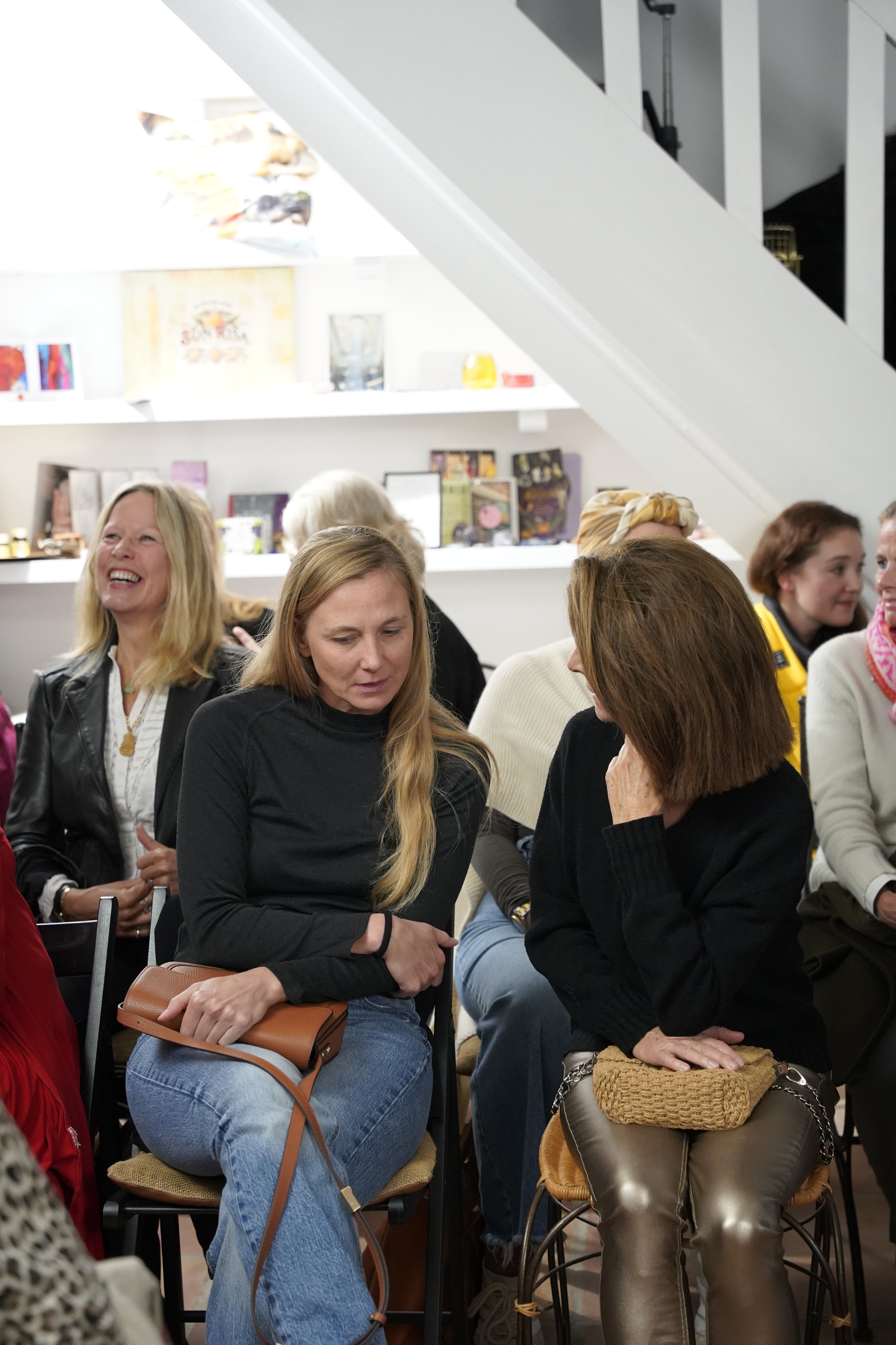 Group of women sitting and talking in a cozy indoor setting, with shelves and artwork on the wall in the background.