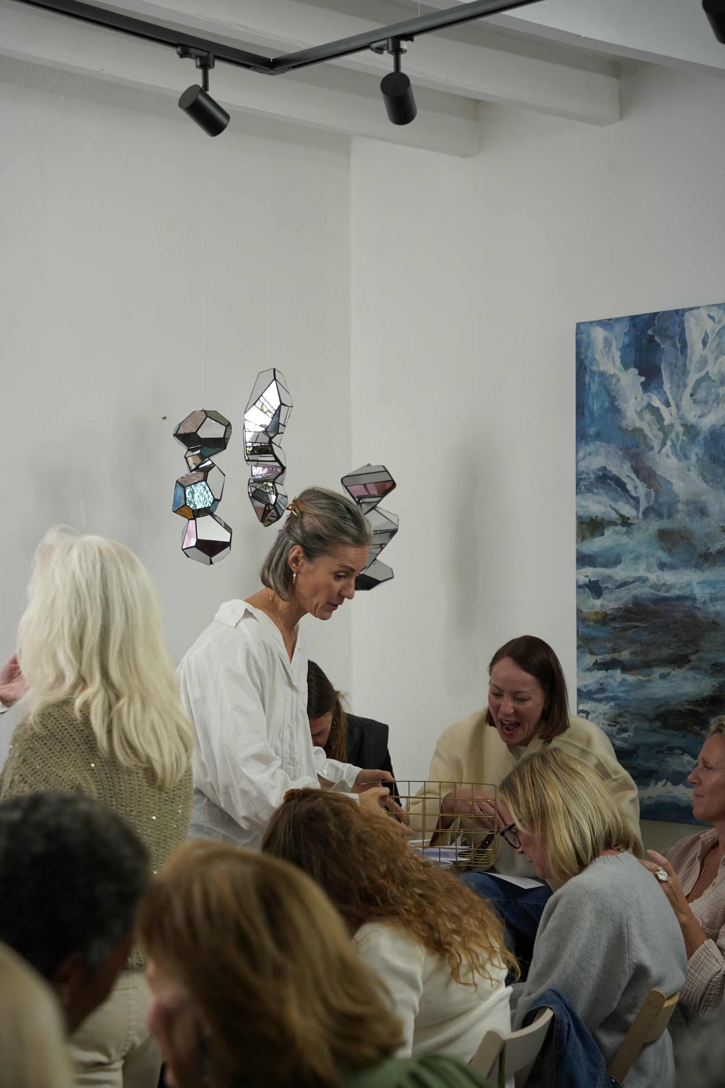 A group of women gathered around a table, engaging in conversation and activities, in an art gallery or similar space with a large blue abstract painting and unique hanging mirror sculptures.