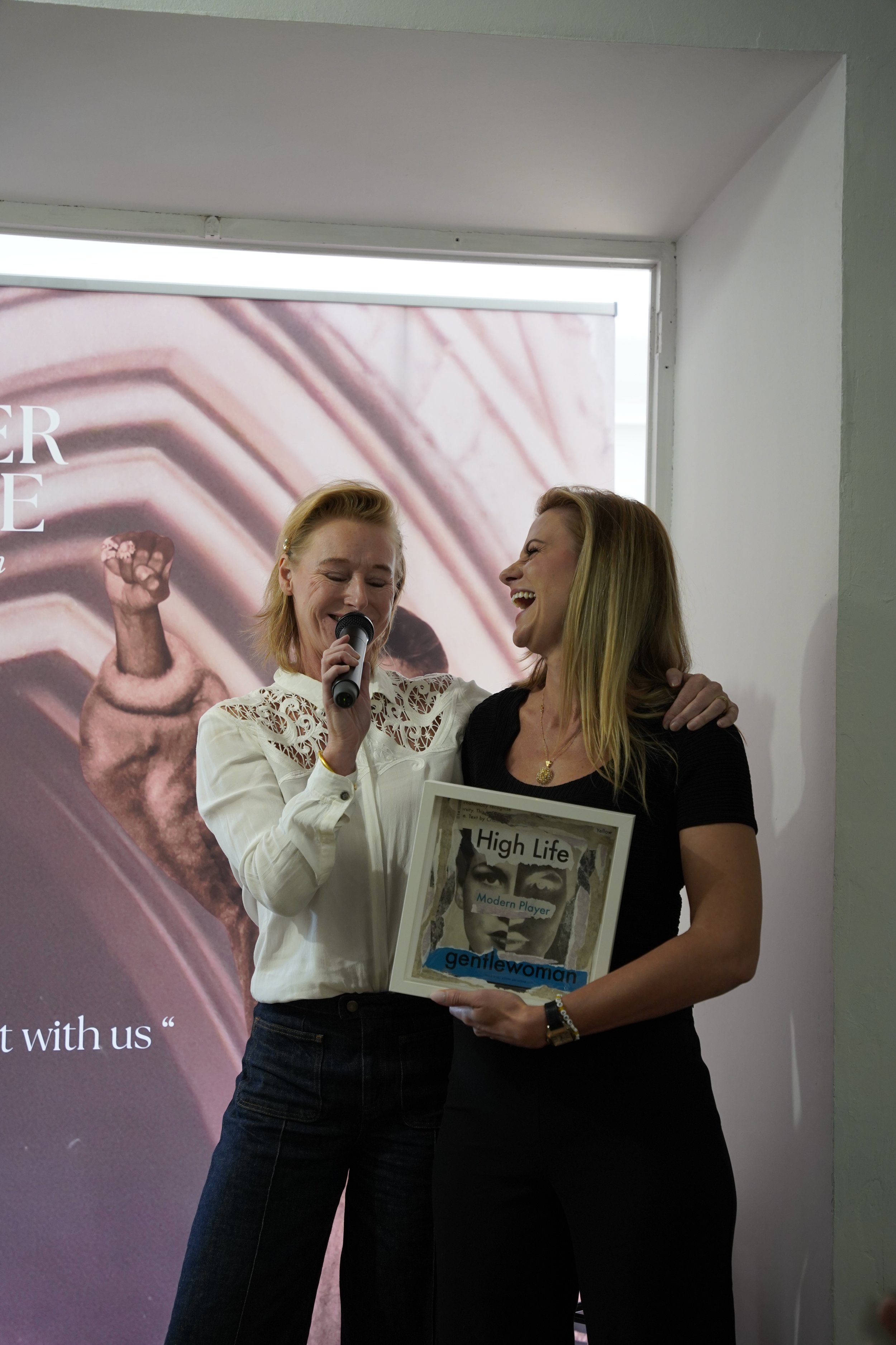 Two women standing close together, smiling and engaging in conversation at an event. One woman holds a microphone and a framed magazine titled 'High Life'. The background features a large poster with part of a person's face and arm, and some visible 