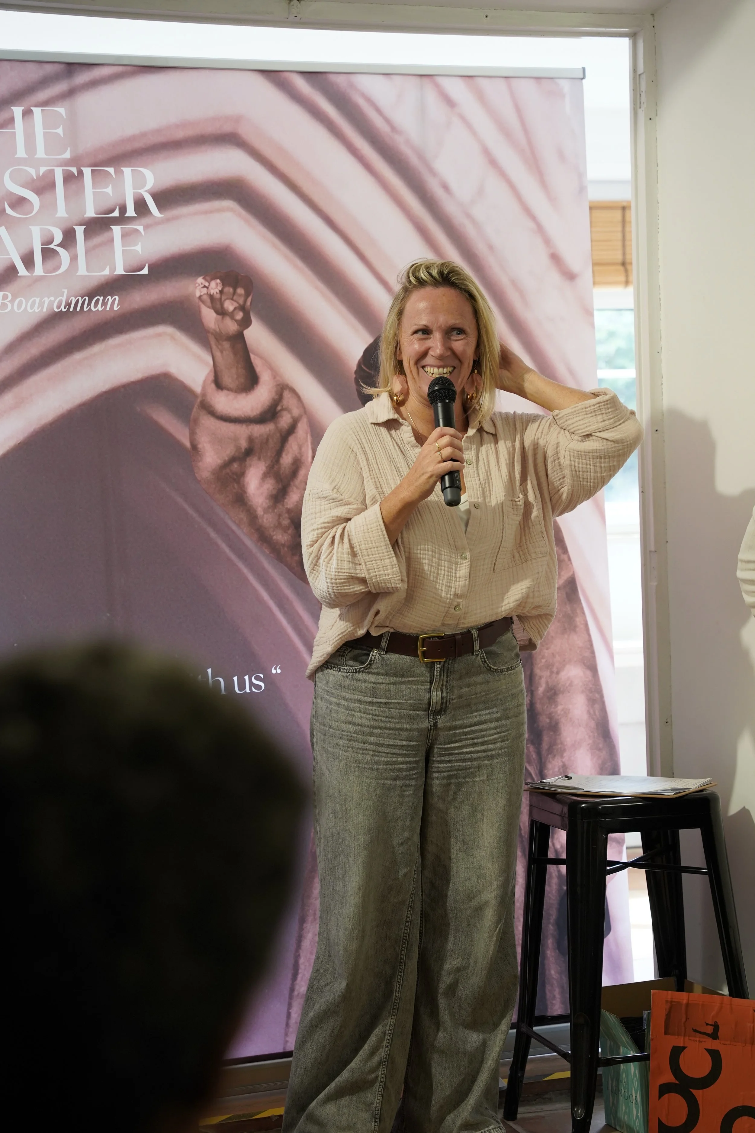 A woman with blonde hair and a beige shirt speaking into a microphone at an event, standing in front of a promotional backdrop that features a raised fist and stylized lines. There are boxes and a stool on the floor nearby.