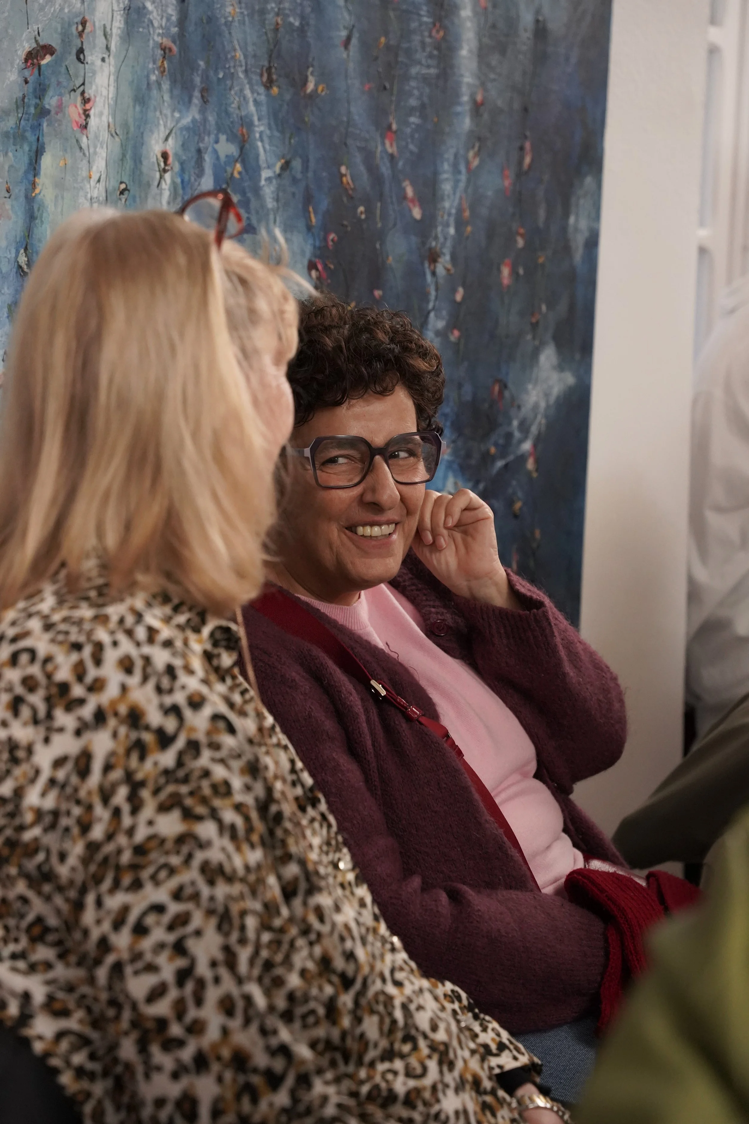 Two women are sitting close together in an indoor setting with a blue abstract painting in the background. One woman has blonde hair and is wearing a leopard print top, while the other has short curly hair, glasses, and is wearing a pink top with a m