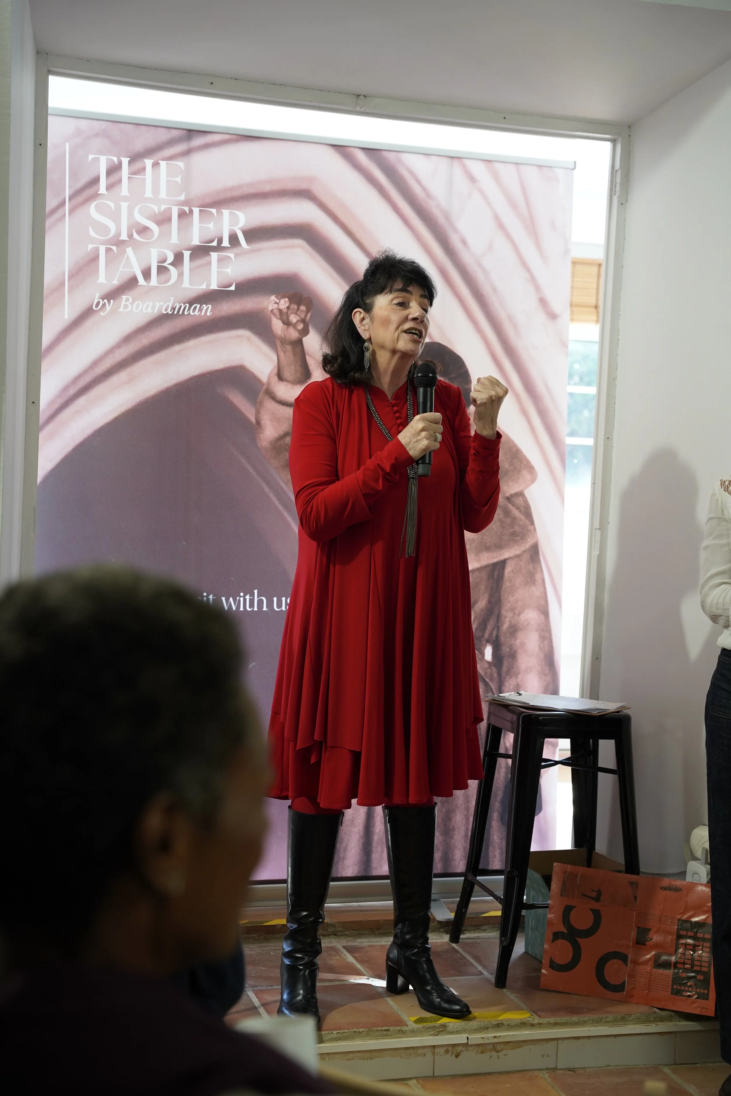 A woman in a red dress and black boots is speaking into a microphone during a presentation. Behind her is a large banner that reads 'The Sister Table by Boardman' with an image of a clenched fist.