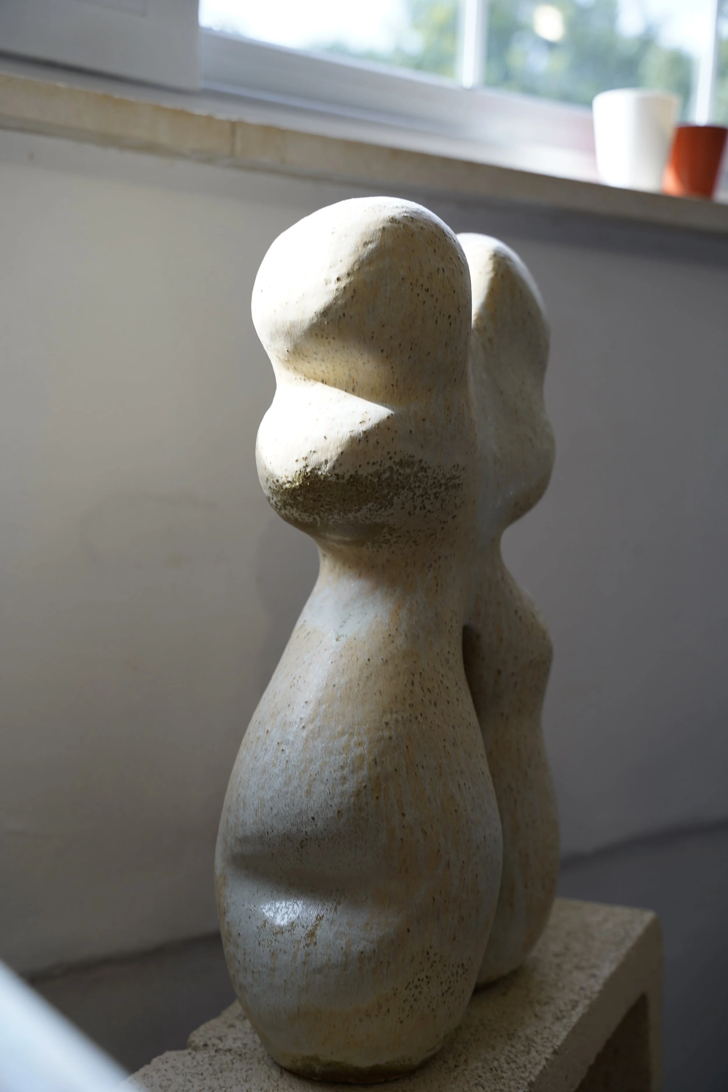 A beige stone sculpture of a stylized female figure with rounded, abstract features, positioned on a rectangular base near a window with blurred planters on the sill in the background.