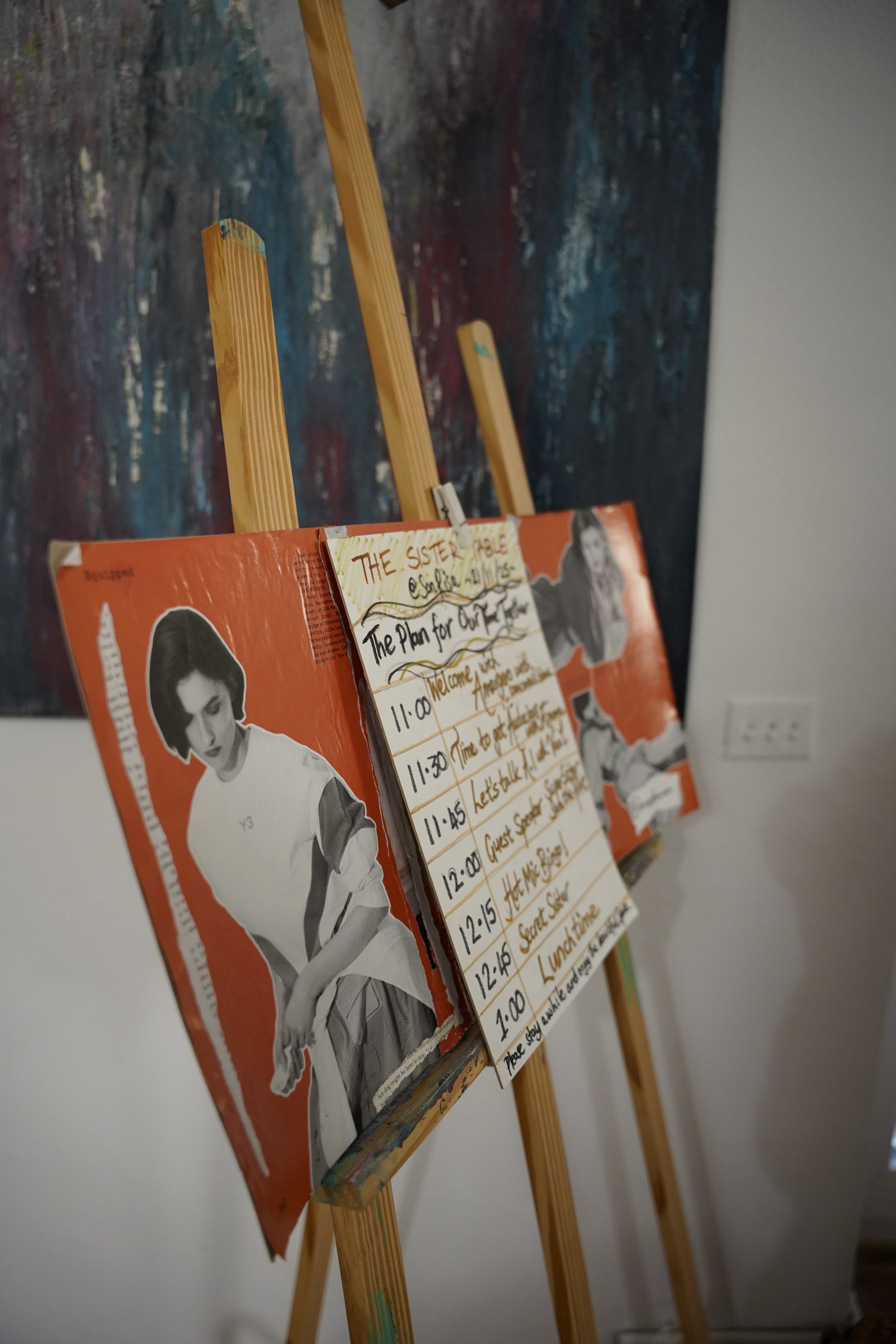 An easel with a laminated poster of a young woman and a handwritten schedule on paper clip. The schedule lists times and events, and the poster appears to be part of an art exhibition or presentation.