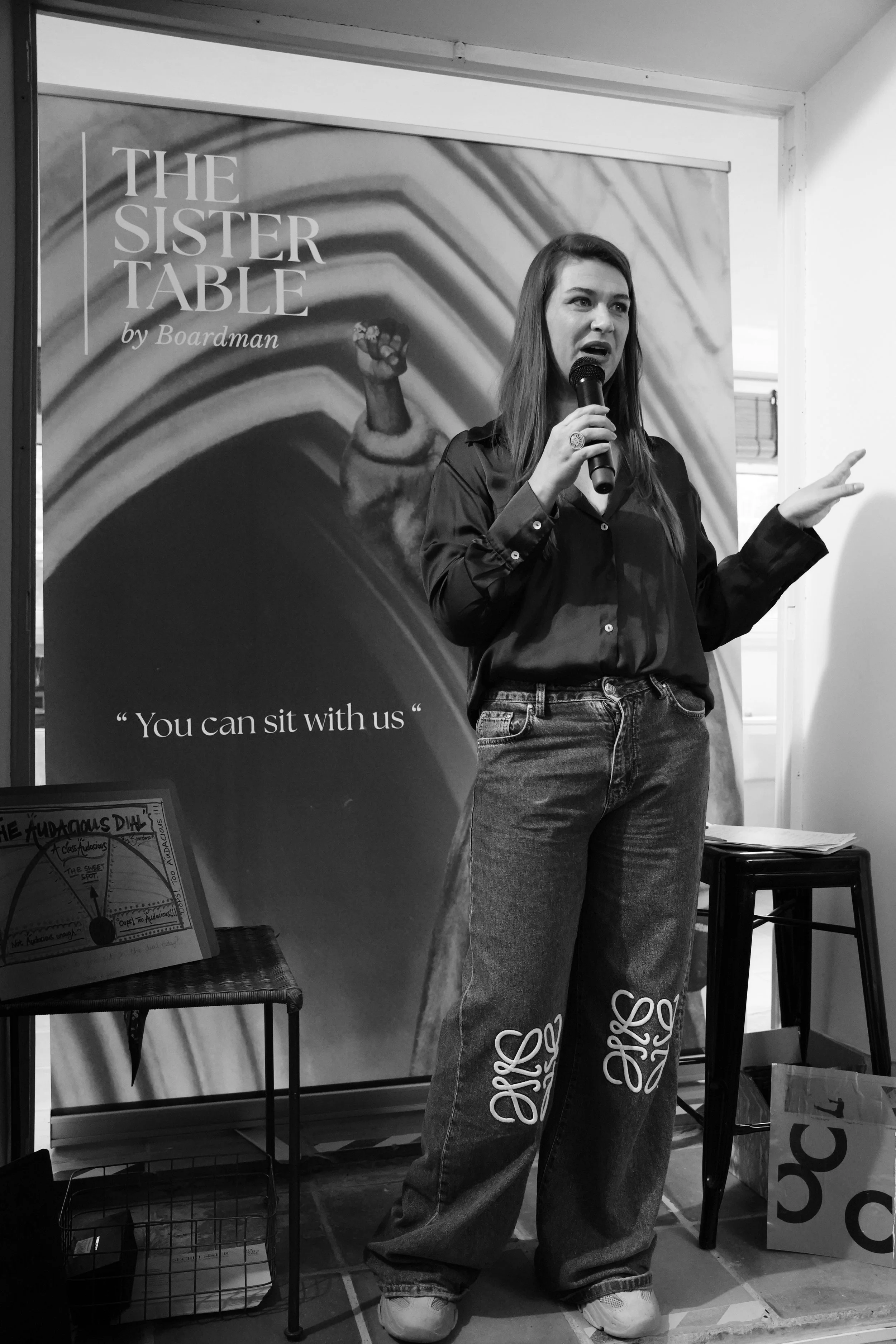 Woman speaking into a microphone at a presentation with a banner behind her that reads 'The Sister Table by Boardman' and a quote 'You can sit with us' at the bottom. There are tables and posters around.