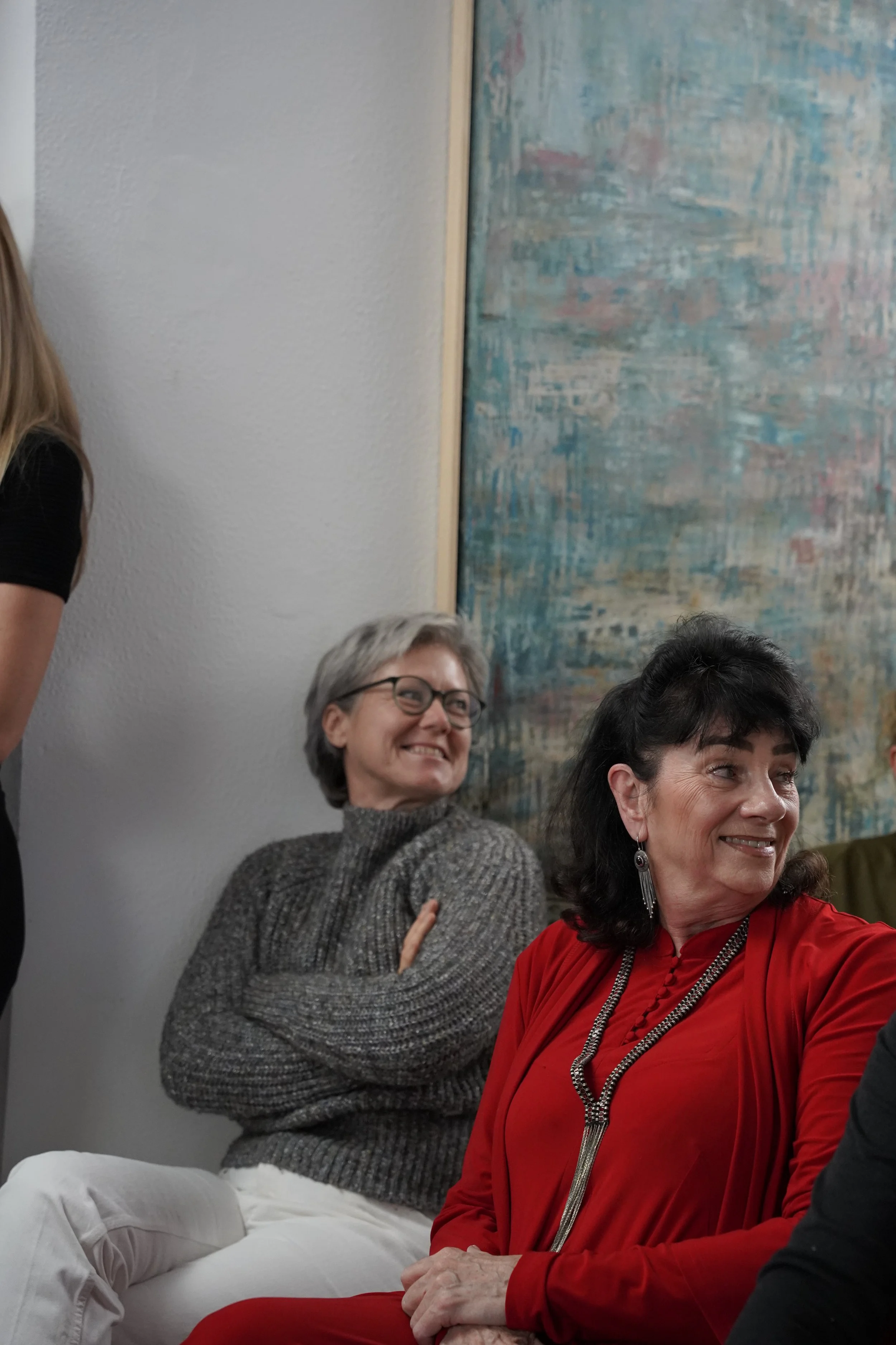 Group of women sitting and smiling at an indoor gathering, with a textured wall and colorful abstract painting in the background.