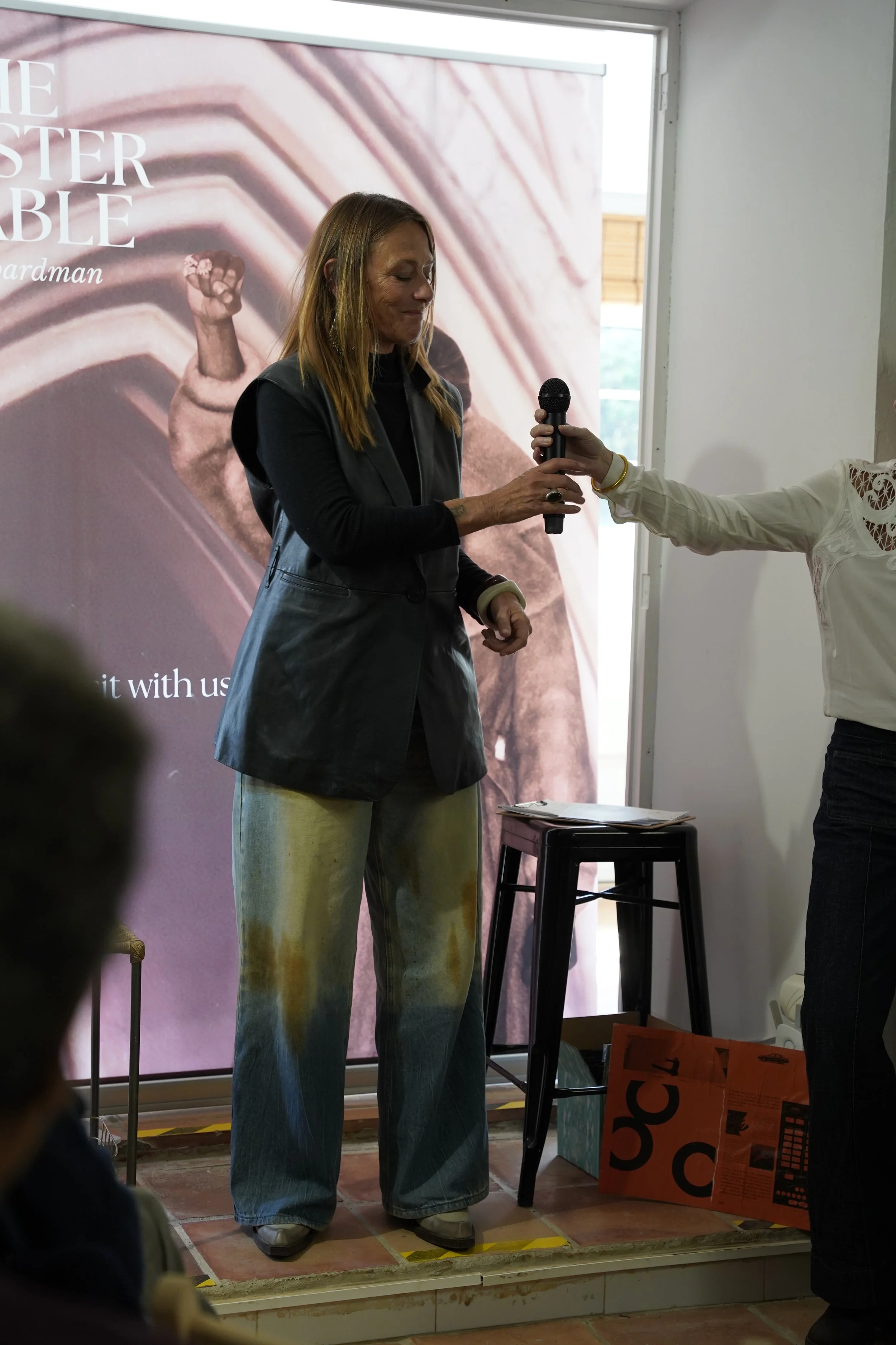 A woman with light brown hair wearing a black vest and jeans stands on a small stage, holding a microphone, with a large poster behind her showing a fist raised in the air and a background of pink and beige abstract patterns.