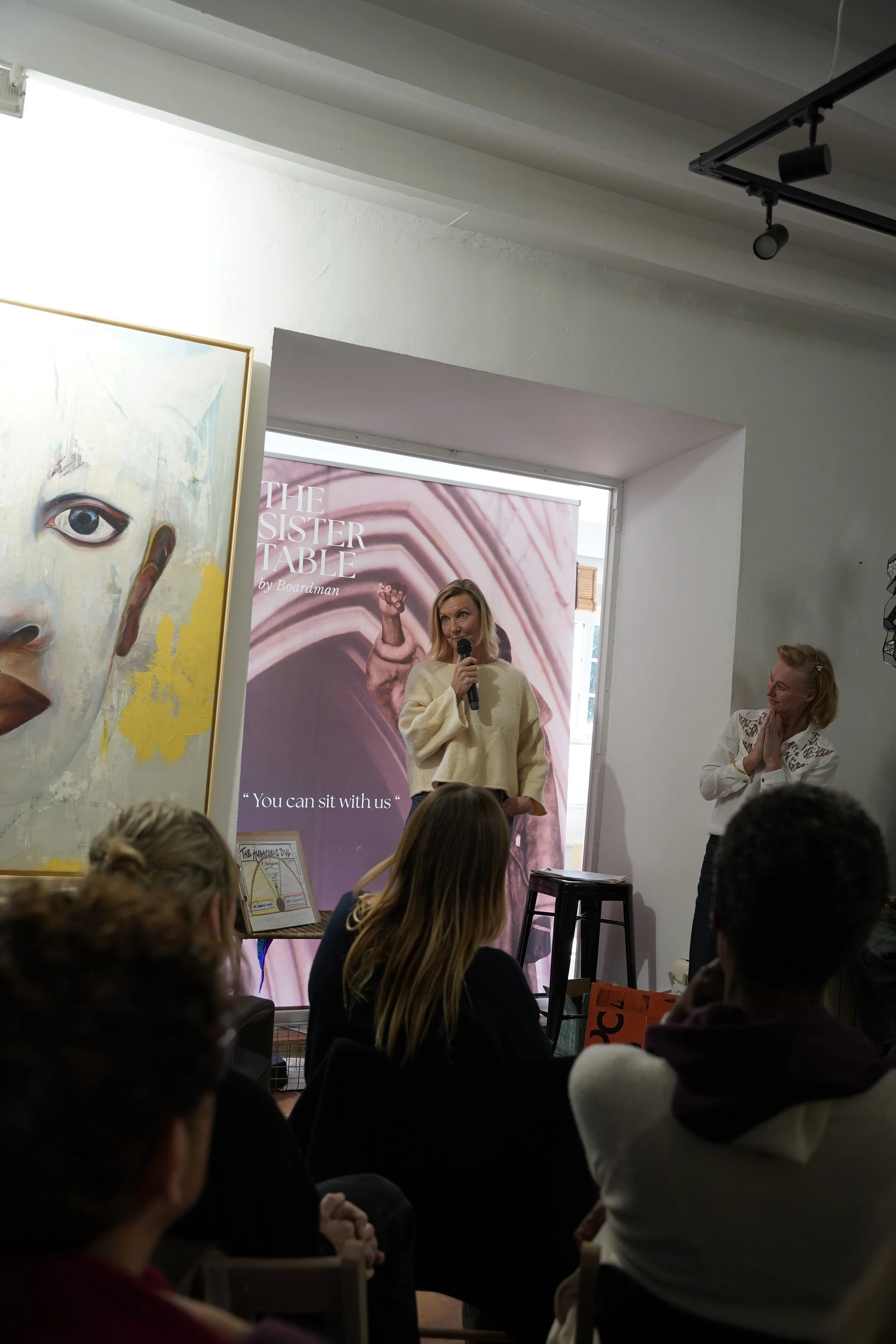 A woman speaking into a microphone at a book event titled 'The Sister Table' by Boardman, with an audience seated facing her. There are artwork and a large window in the background.
