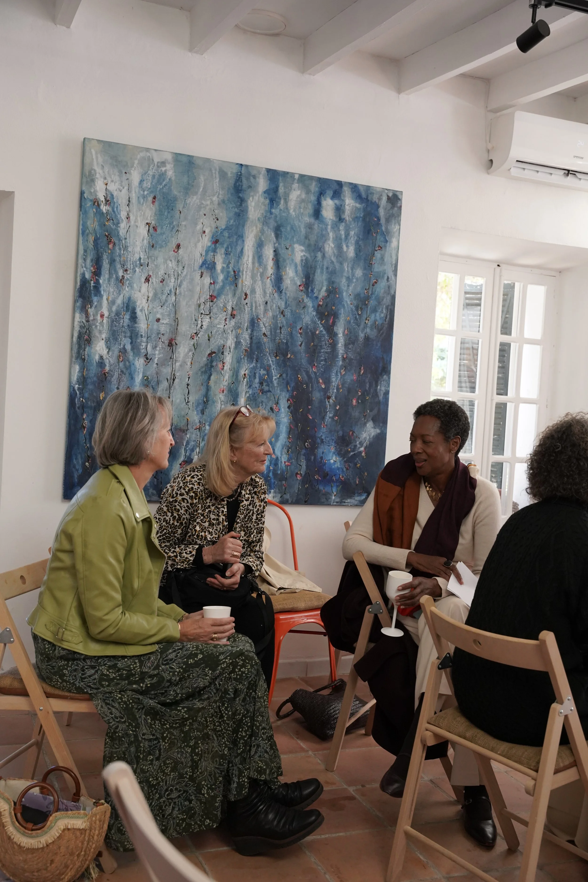 A group of women sitting and chatting in a bright room with white walls, wooden beams, and a large abstract blue painting on the wall. Some women are holding cups.