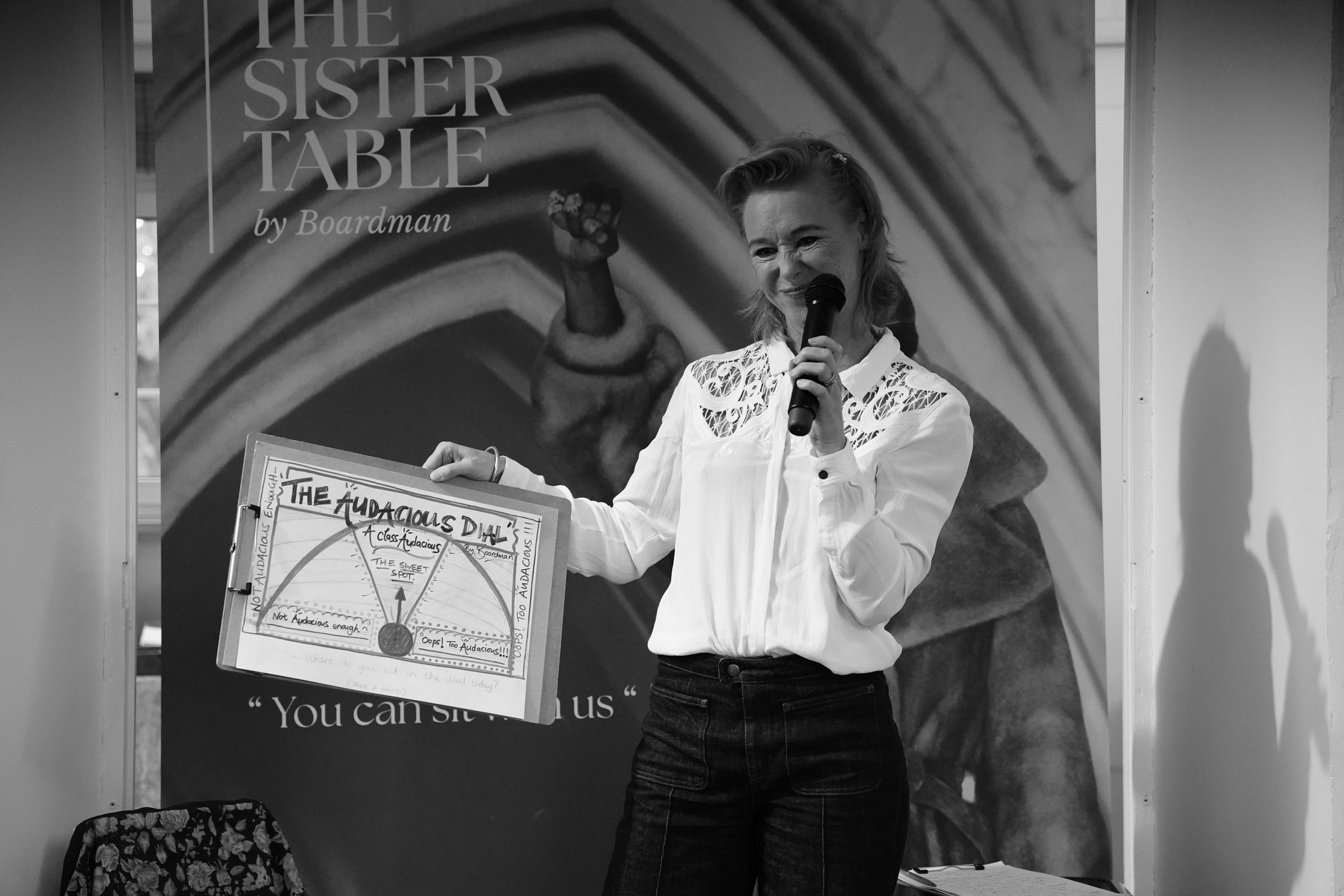 A woman holding a microphone and a poster stands in front of a large poster that reads 'The Sister Table by Boardman'. The woman is wearing a white blouse with lace details and dark pants, and is gesturing with her right hand. The poster behind her c