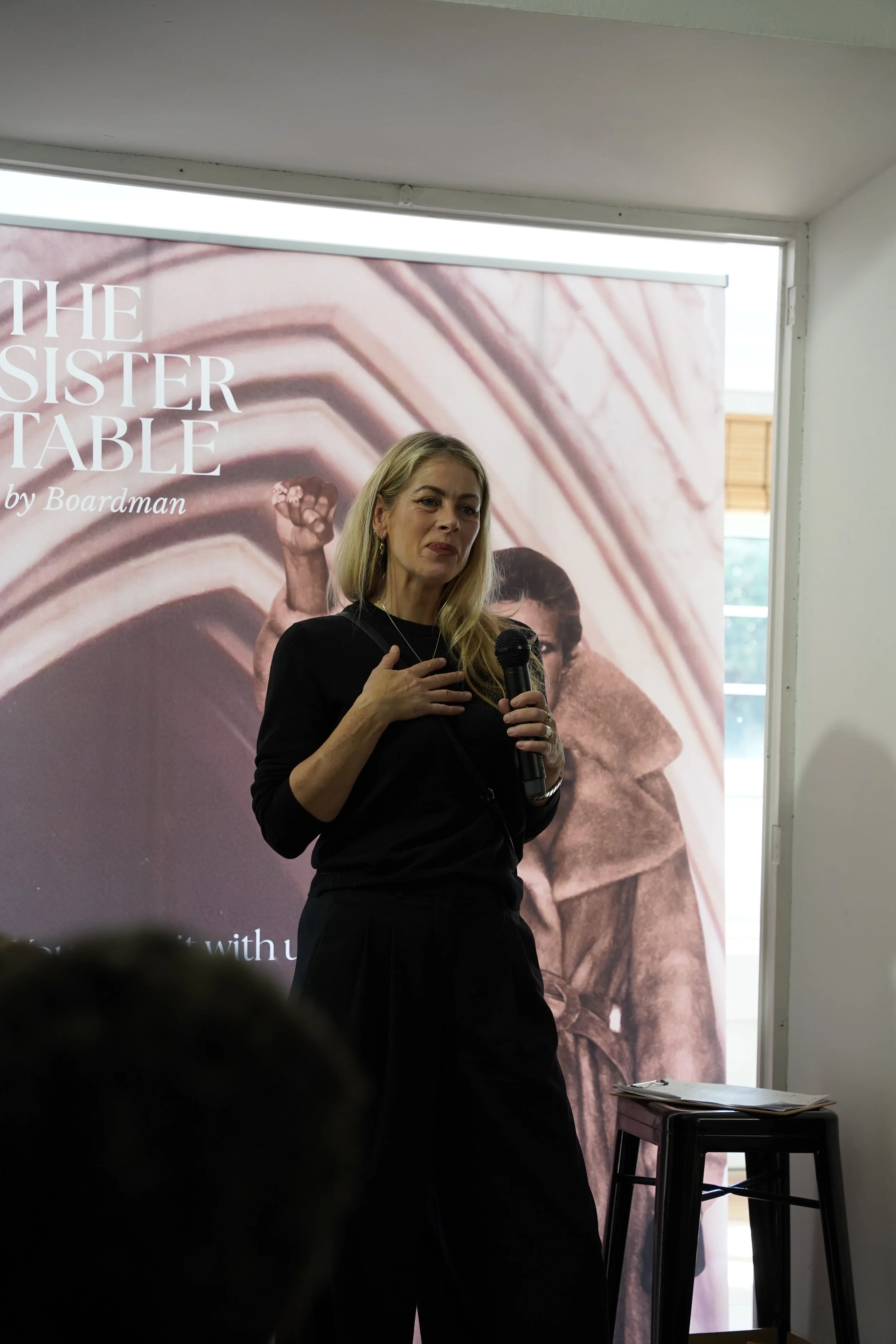 A woman holding a microphone standing in front of a promotional poster for the book 'The Sister Table' by Boardman. The poster features a close-up image of a fanned stack of pinkish-brown plates, with a partial image of a woman's face.