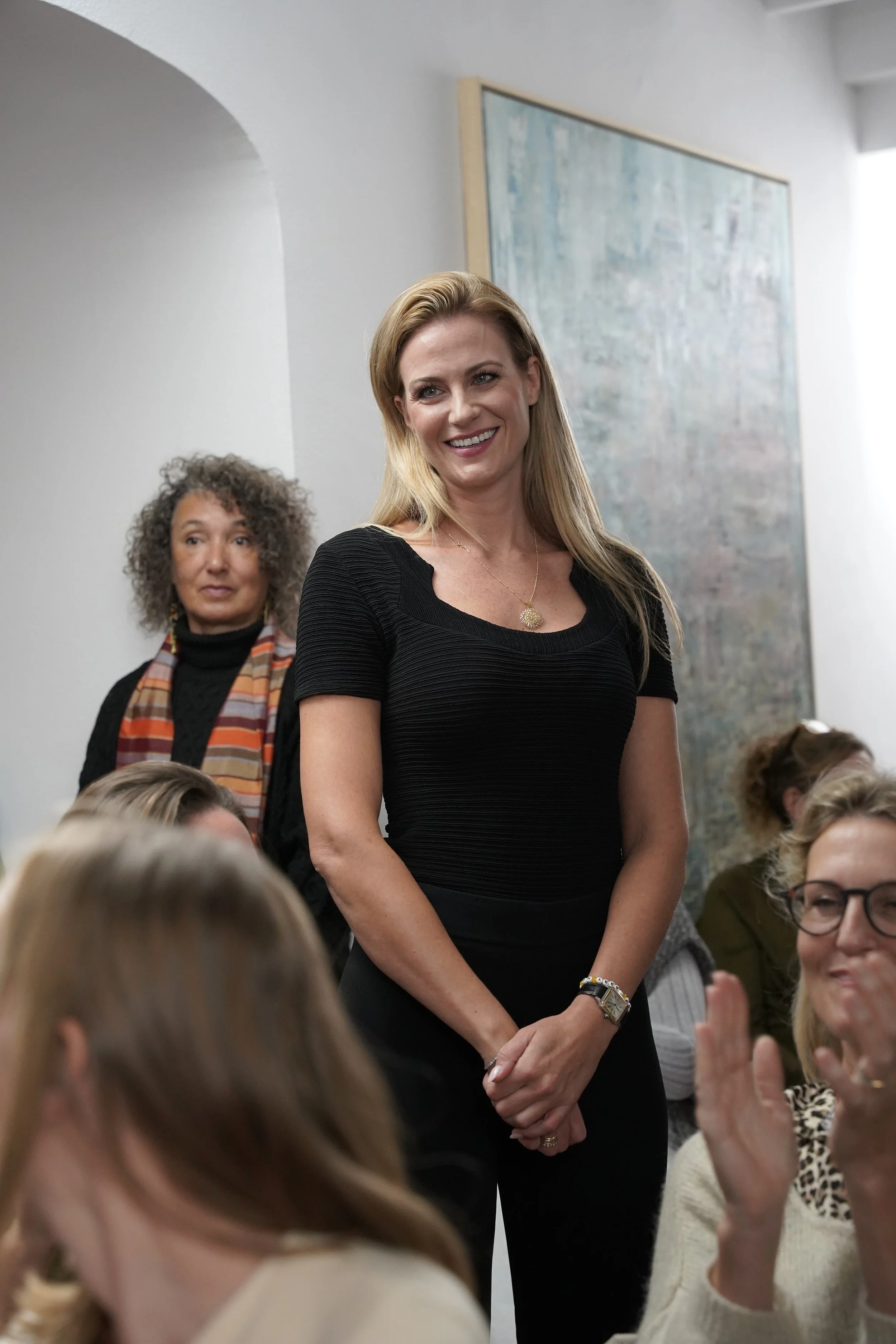 A woman with blonde hair smiling, wearing a black dress and necklace, standing among a group of seated women in a room with white walls and abstract art.