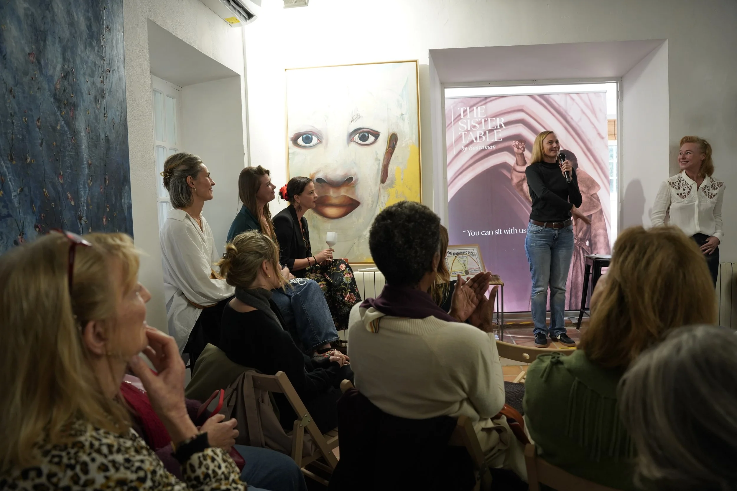 A group of women attending a presentation or workshop in an art gallery or similar setting. One woman with a microphone stands in front of a pink banner that reads 'The Sister Table by Boardman'. The audience is seated and listening, some clapping. T
