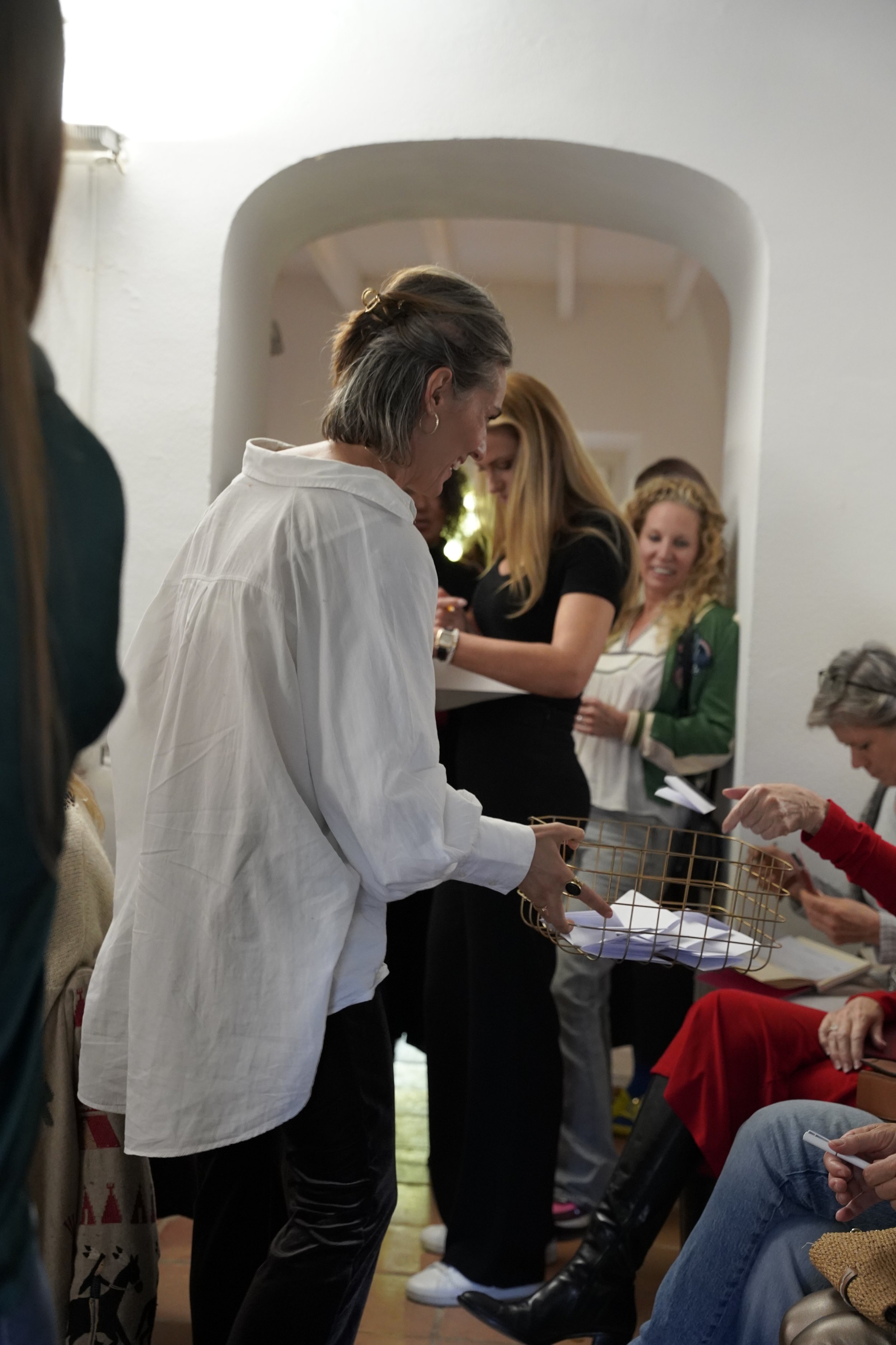 A woman in a white shirt standing and looking down at papers or objects in a wire basket on a table. Other people are seated and standing around her, some looking at papers or items. The setting appears to be indoors with white walls and an archway.