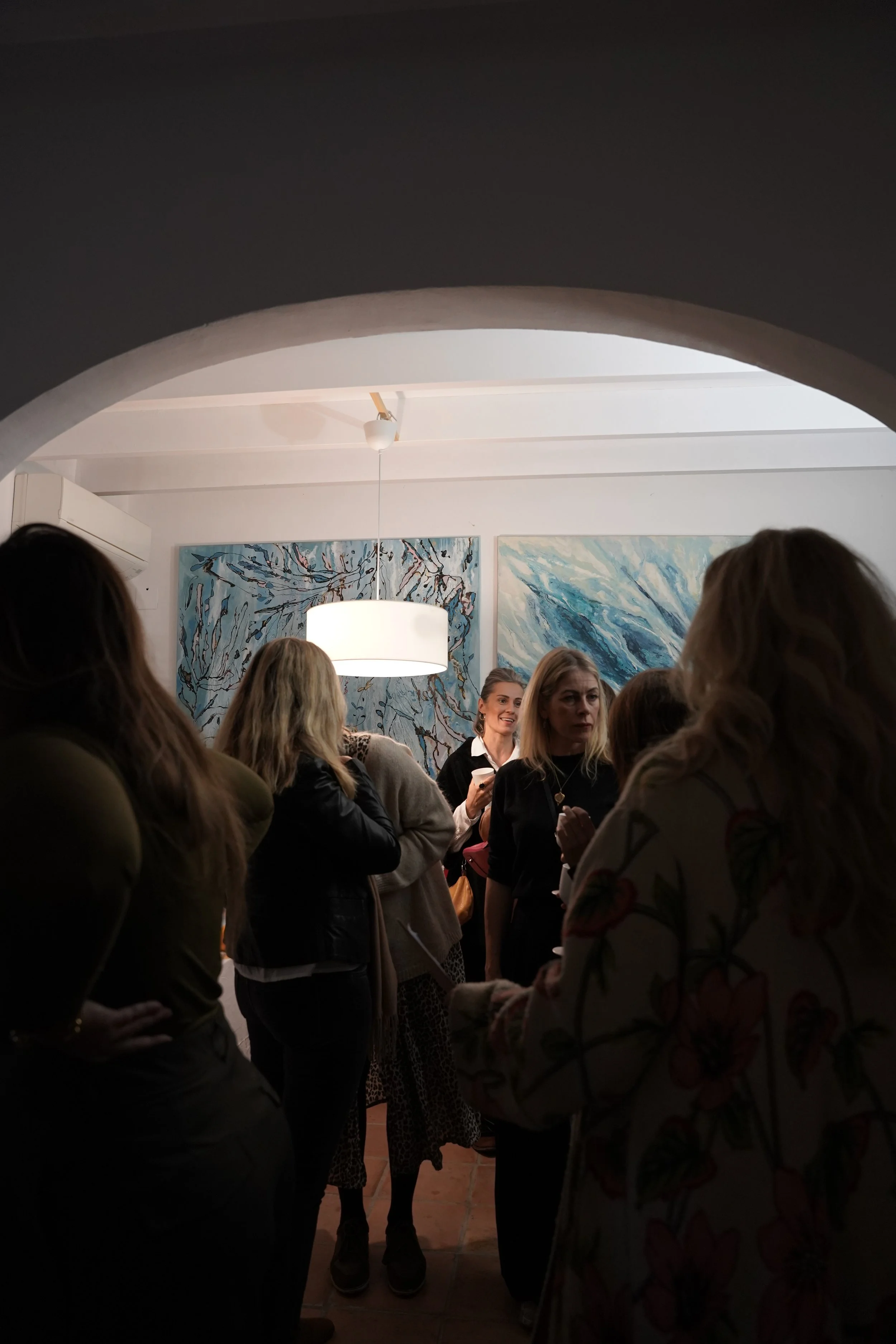 A group of women gathered in an art gallery with abstract paintings on the wall, some wearing patterned clothing, under a ceiling with pendant lighting.
