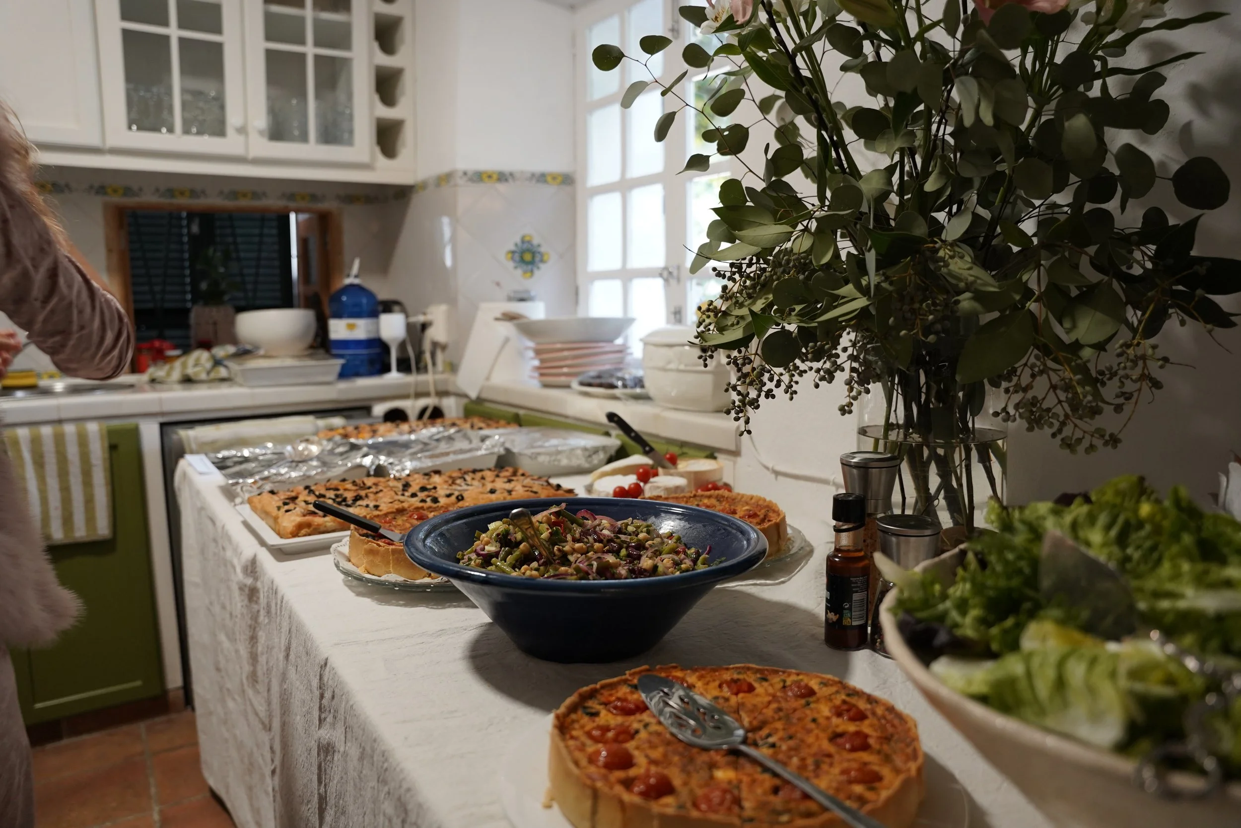 A kitchen countertop with various dishes including a pizza, a salad, and other foods, with a large bouquet of green leafy plants on the right side.