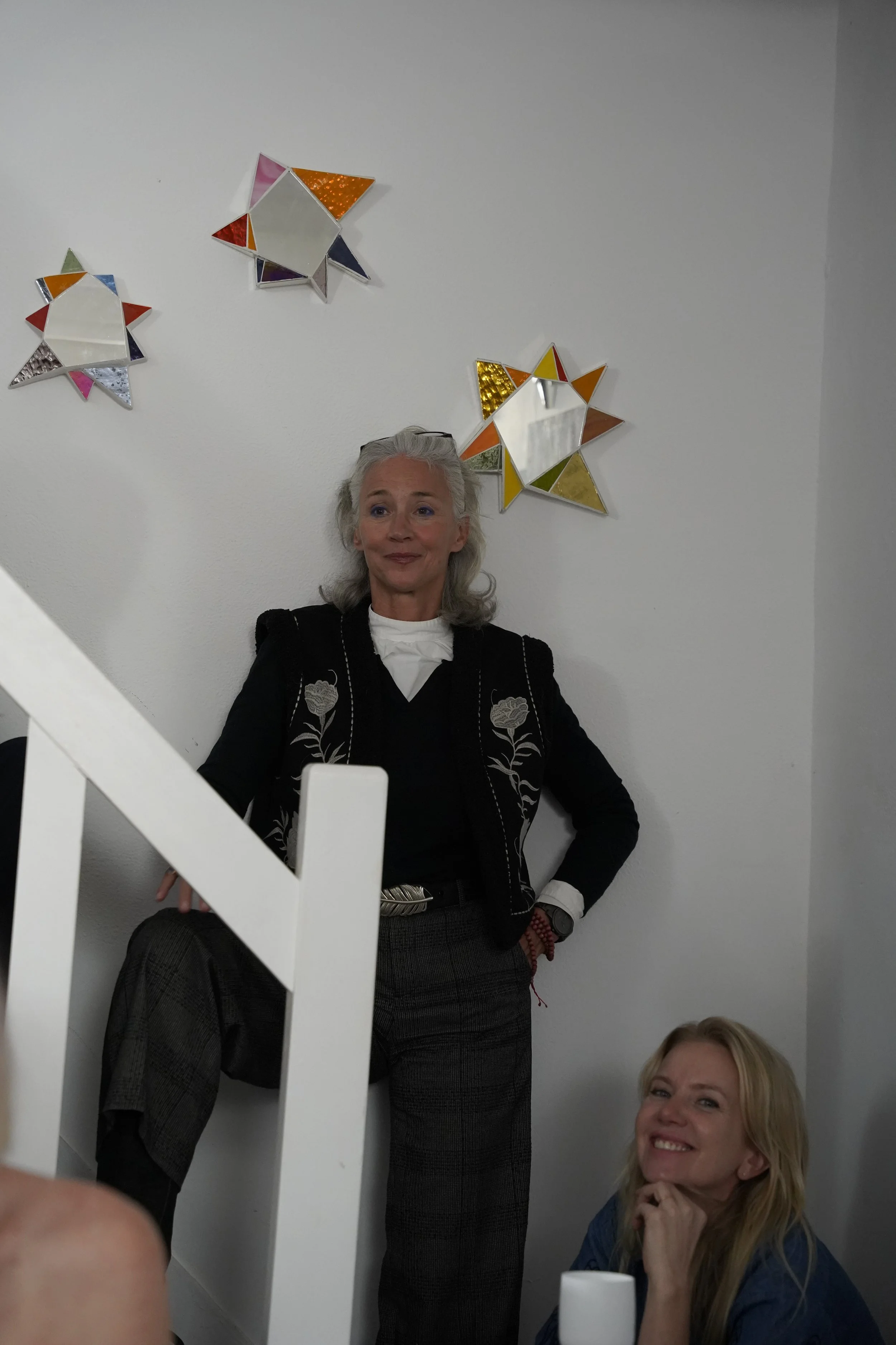 Two women indoors in front of a white wall decorated with three star-shaped mirrors with colored borders. One woman with gray hair, dressed in a black jacket and gray plaid pants, is standing with one leg resting on a step and her hand on her waist. 