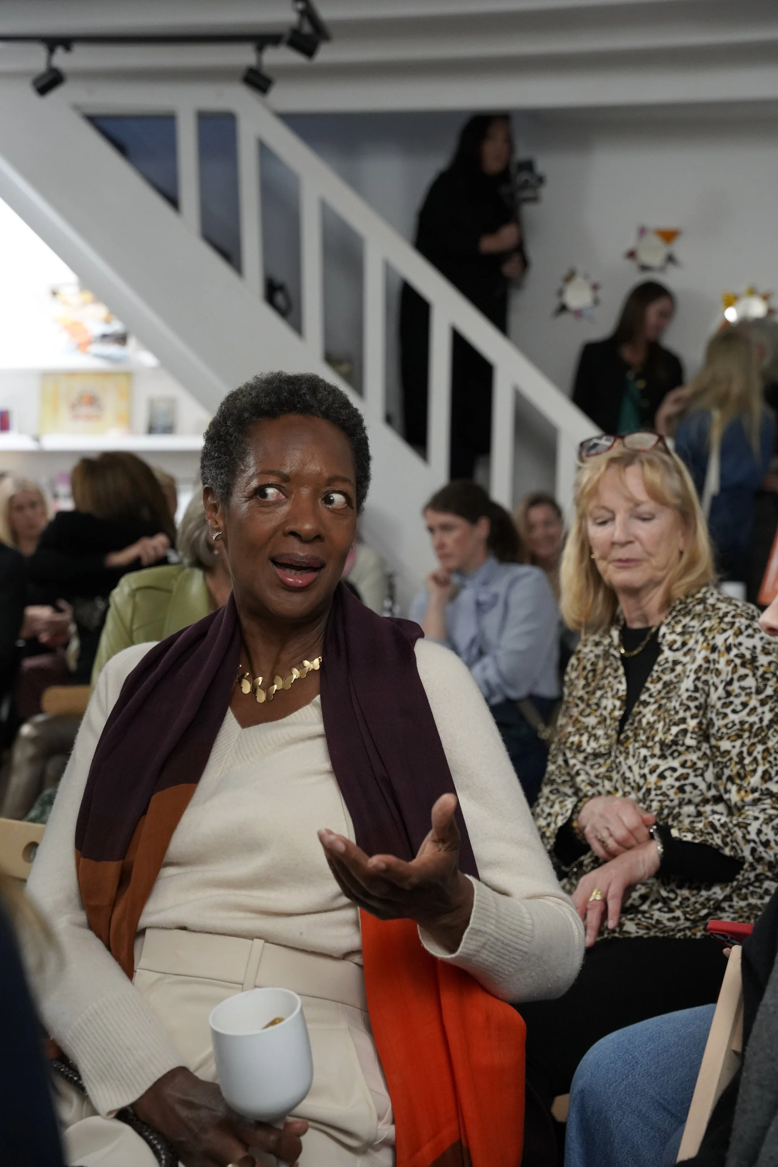A group of women gathered indoors at a social event, with one woman in the foreground speaking and holding a white mug, while others are engaged in conversation in the background.