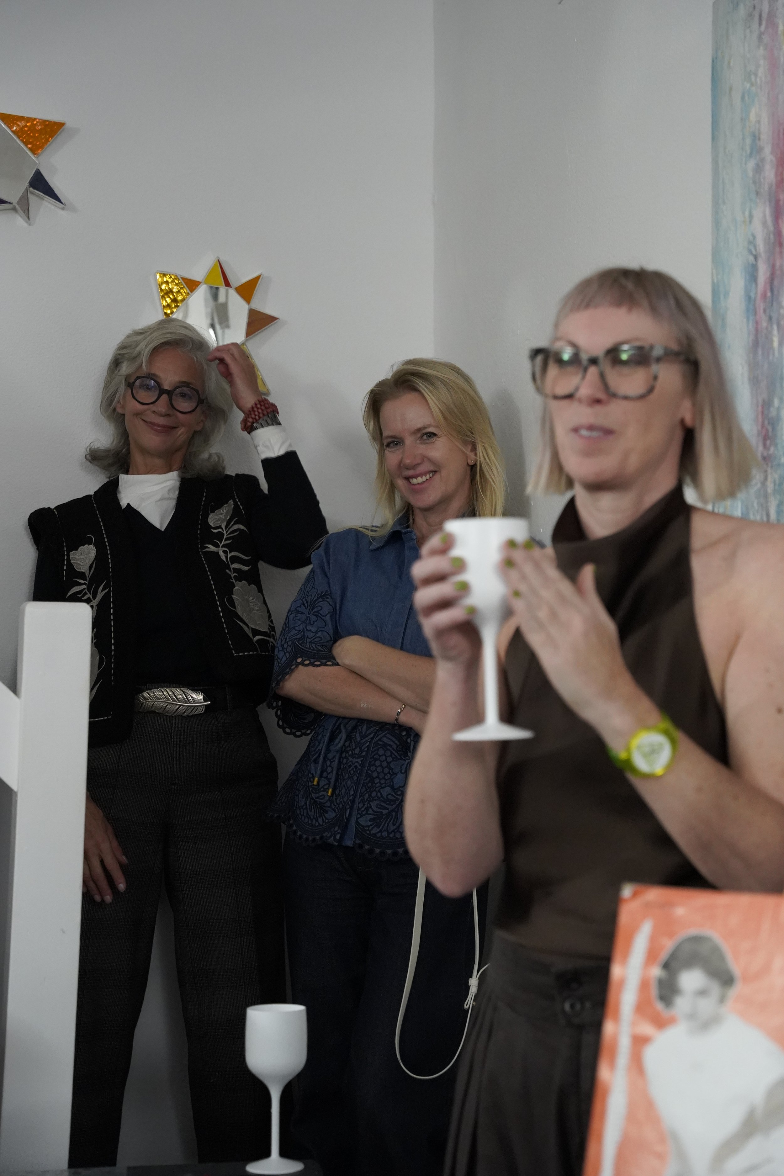 Three women standing indoors, one with gray hair and glasses touching her hair, another with blonde hair smiling, and the third with blonde hair and glasses holding a white wine glass, with colorful star-shaped decorations on the wall behind them.