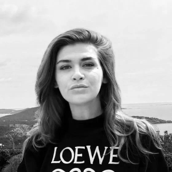 Black and white photo of a woman with long hair, wearing a black shirt with the words "LOEWE" visible, outdoors with a landscape background.
