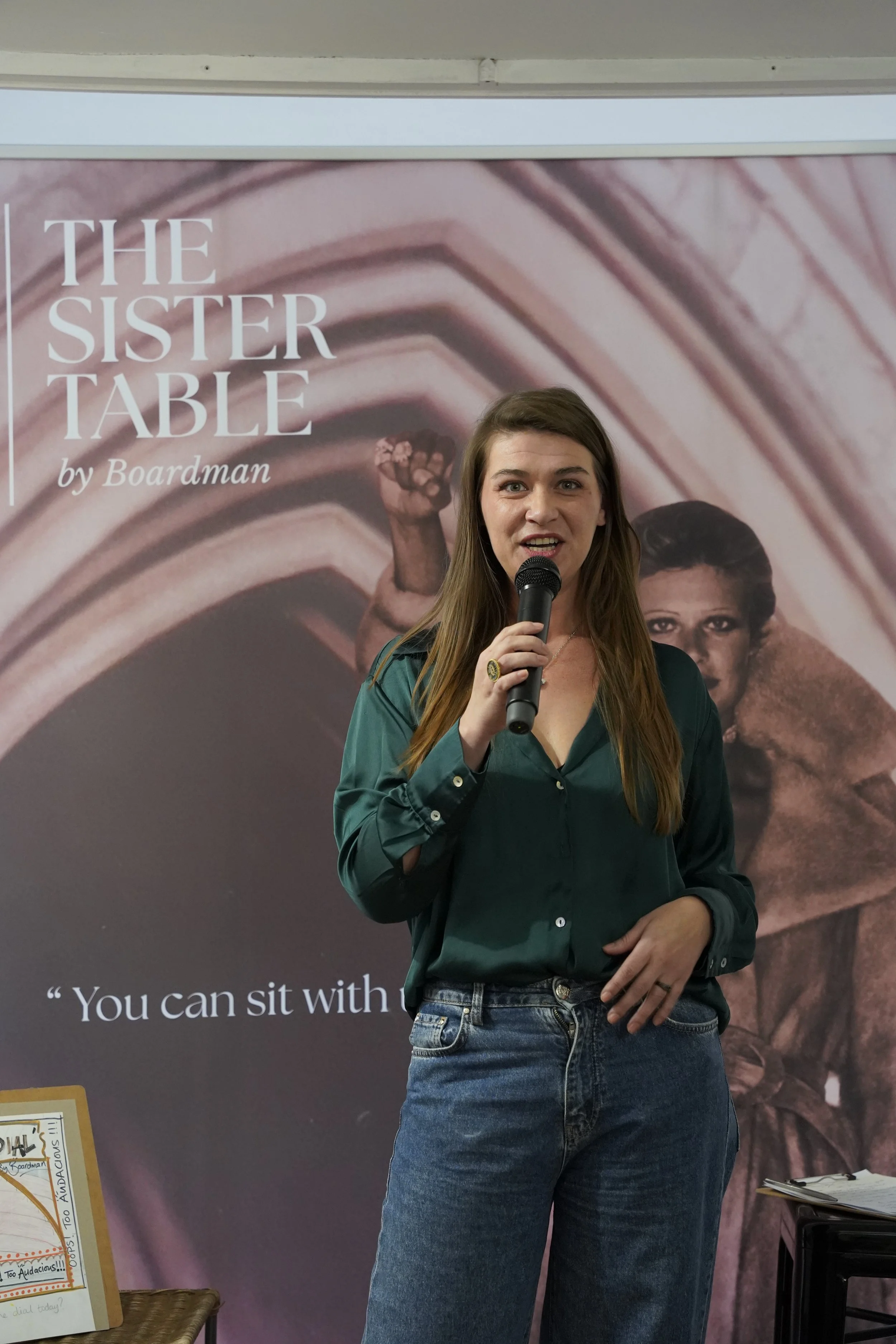 A woman with long brown hair holding a microphone and speaking in front of a large poster. She is wearing a dark green shirt and blue jeans. The poster behind her has the title 'The Sister Table' by Boardman and features an image of a woman with her 