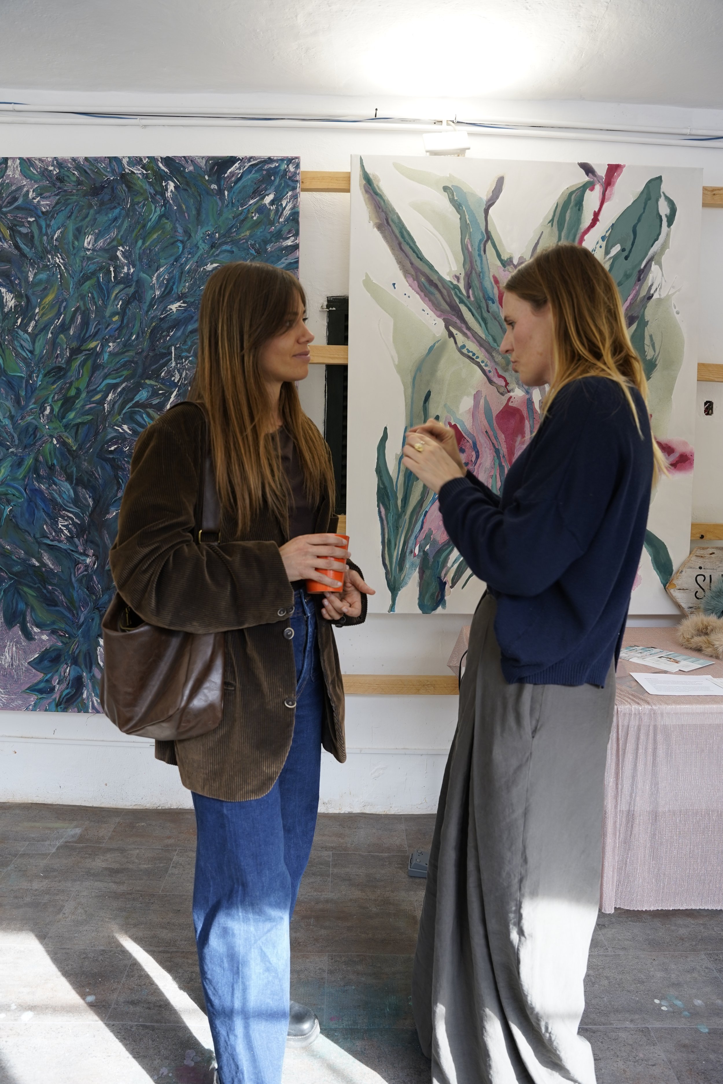 Two women engaged in conversation at an art gallery space, with abstract floral paintings displayed on the wall behind them.
