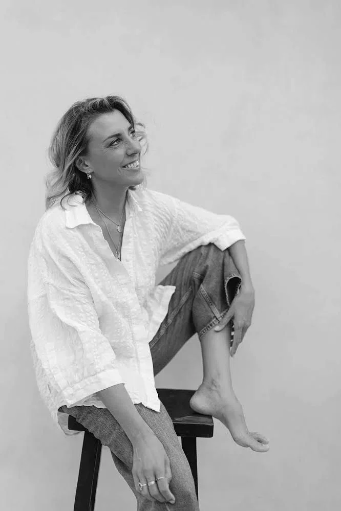 A woman smiling and sitting on a chair with one leg resting on the seat, wearing a white shirt and jeans, against a plain background.