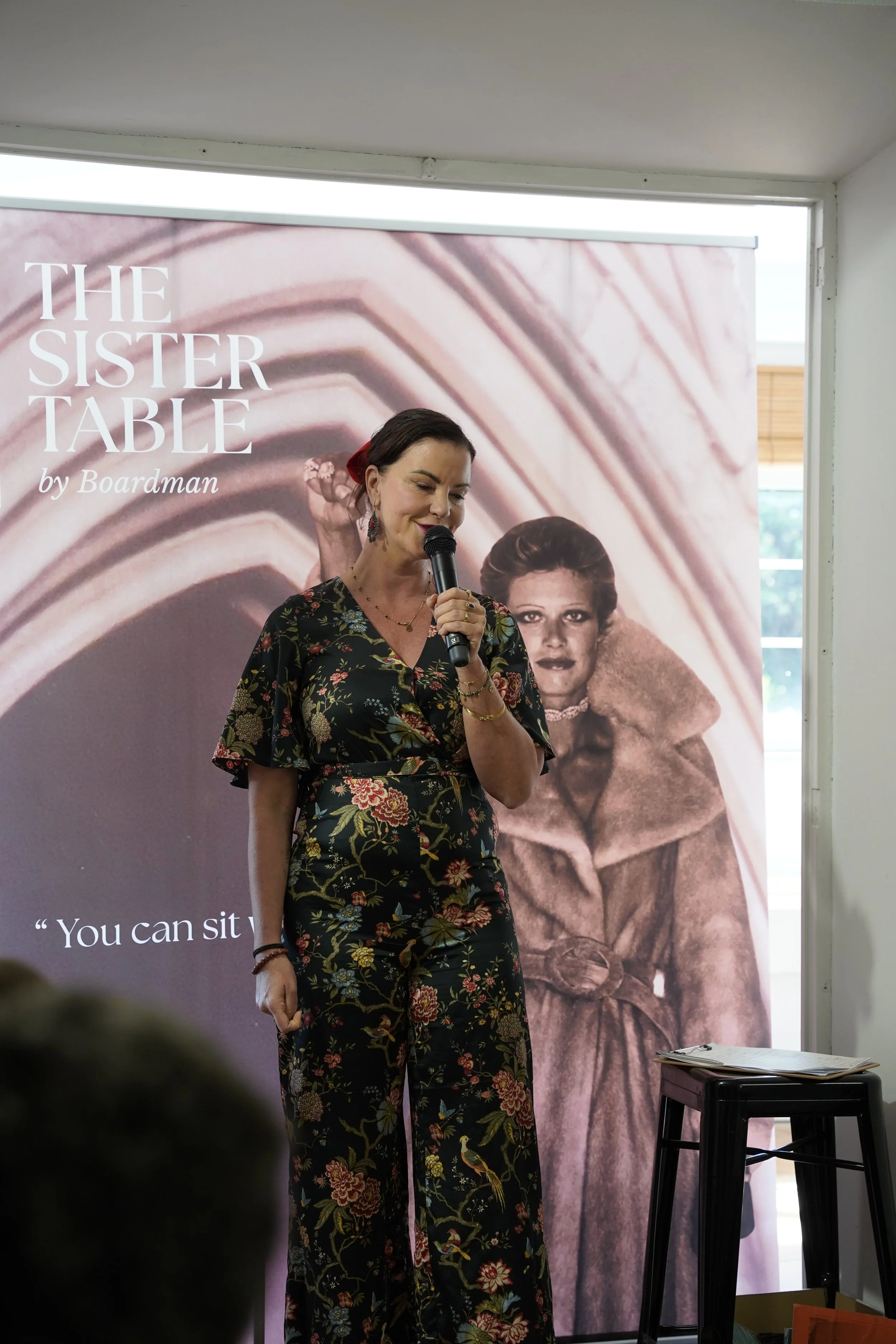 A woman with dark hair and earrings holding a microphone, wearing a black floral jumpsuit, standing in front of a large poster with a woman and architectural details. The poster reads 'THE SISTER TABLE by Boardman'.