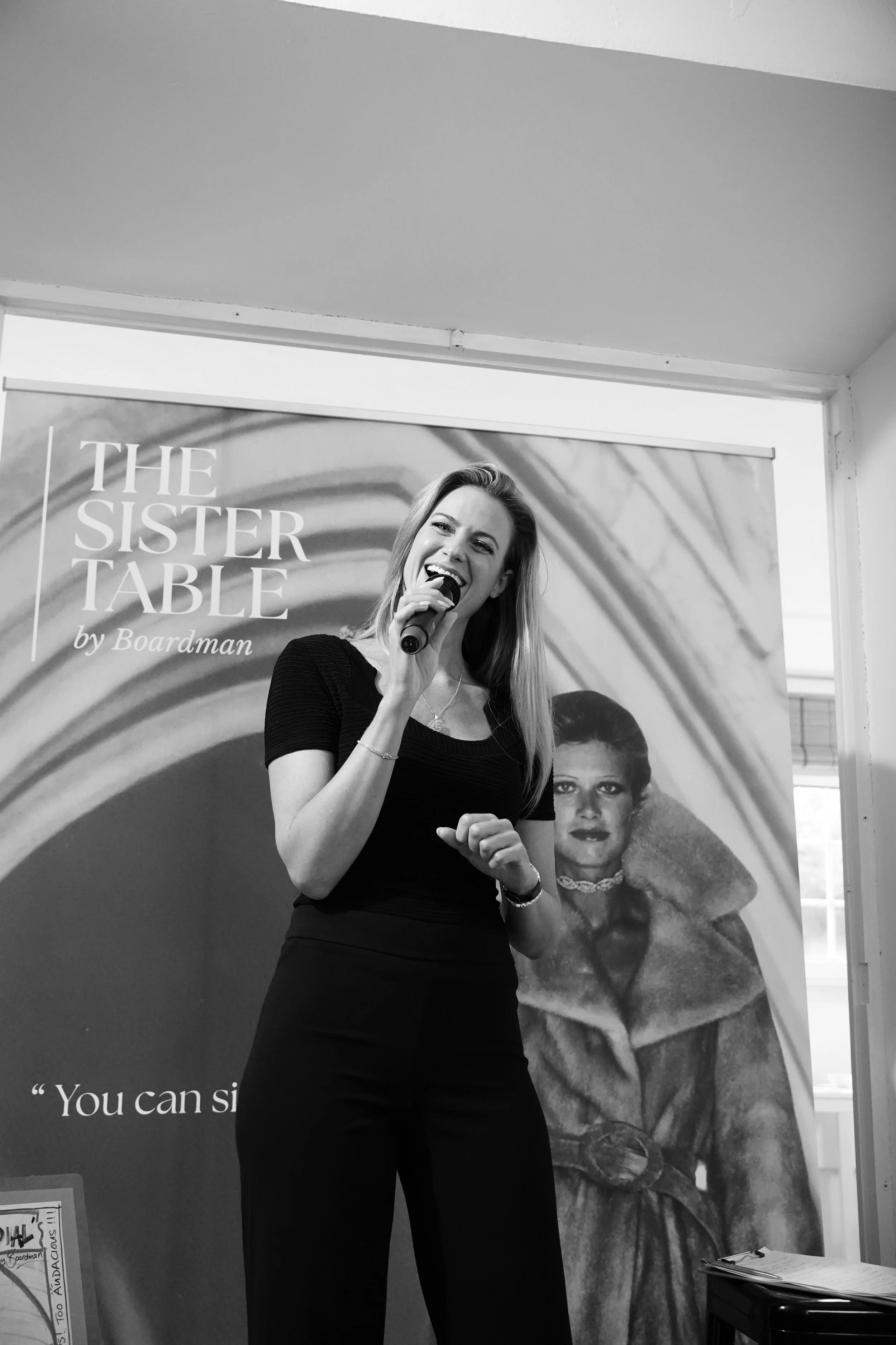 A woman with long hair speaking into a microphone at an event, standing in front of a promotional poster featuring the title 'THE SISTER TABLE by Boardman' and a woman with short hair and fur clothing in the background.