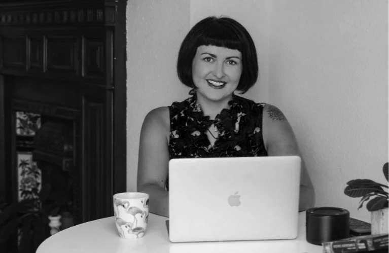 A woman with short dark hair and a patterned sleeveless top sits at a table with a laptop, a mug, and a plant, smiling at the camera in a home setting.
