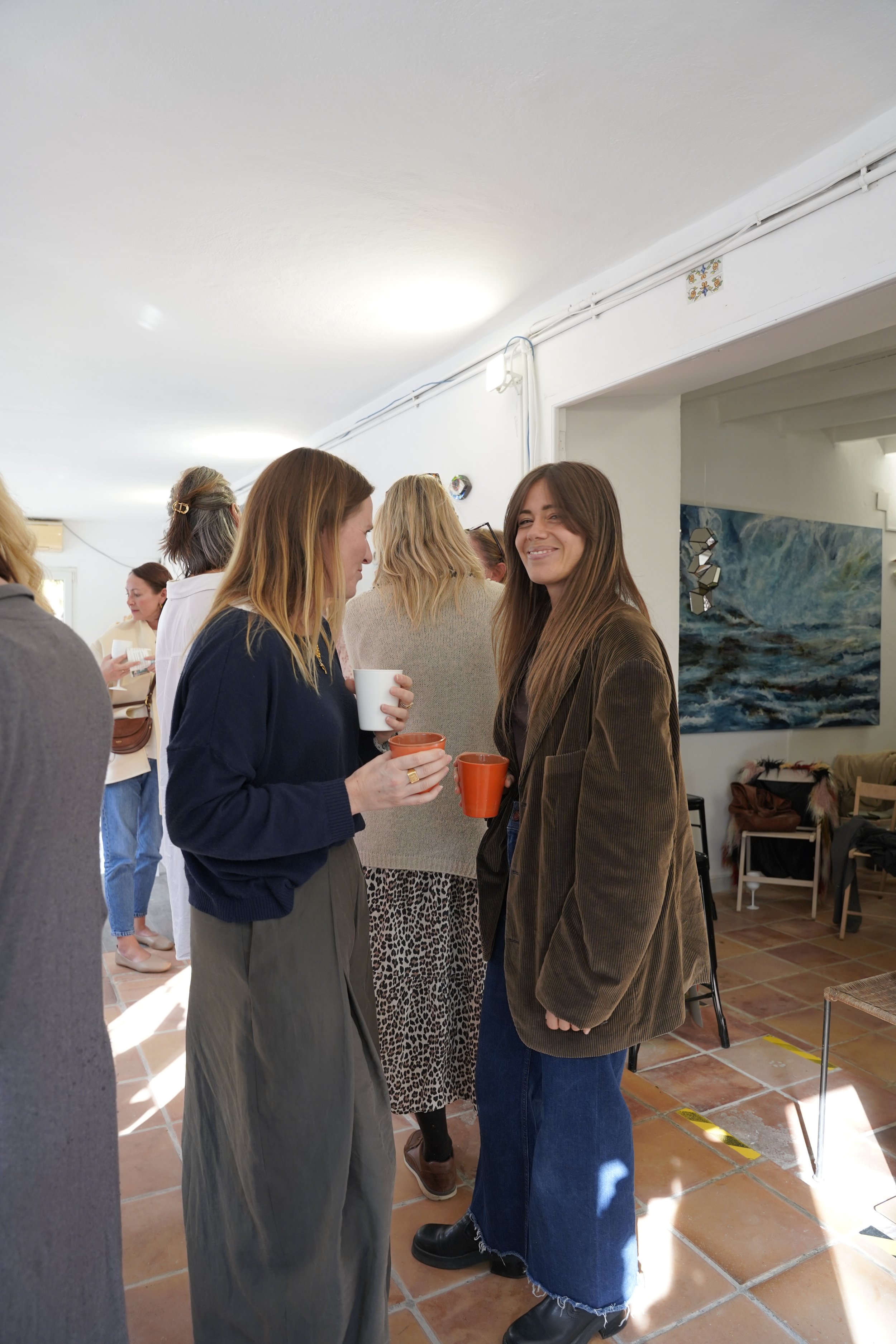 A group of women at an indoor social gathering, engaging in conversation and holding cups. The setting has white walls, artwork, and a tiled floor, with bright natural light streaming in.