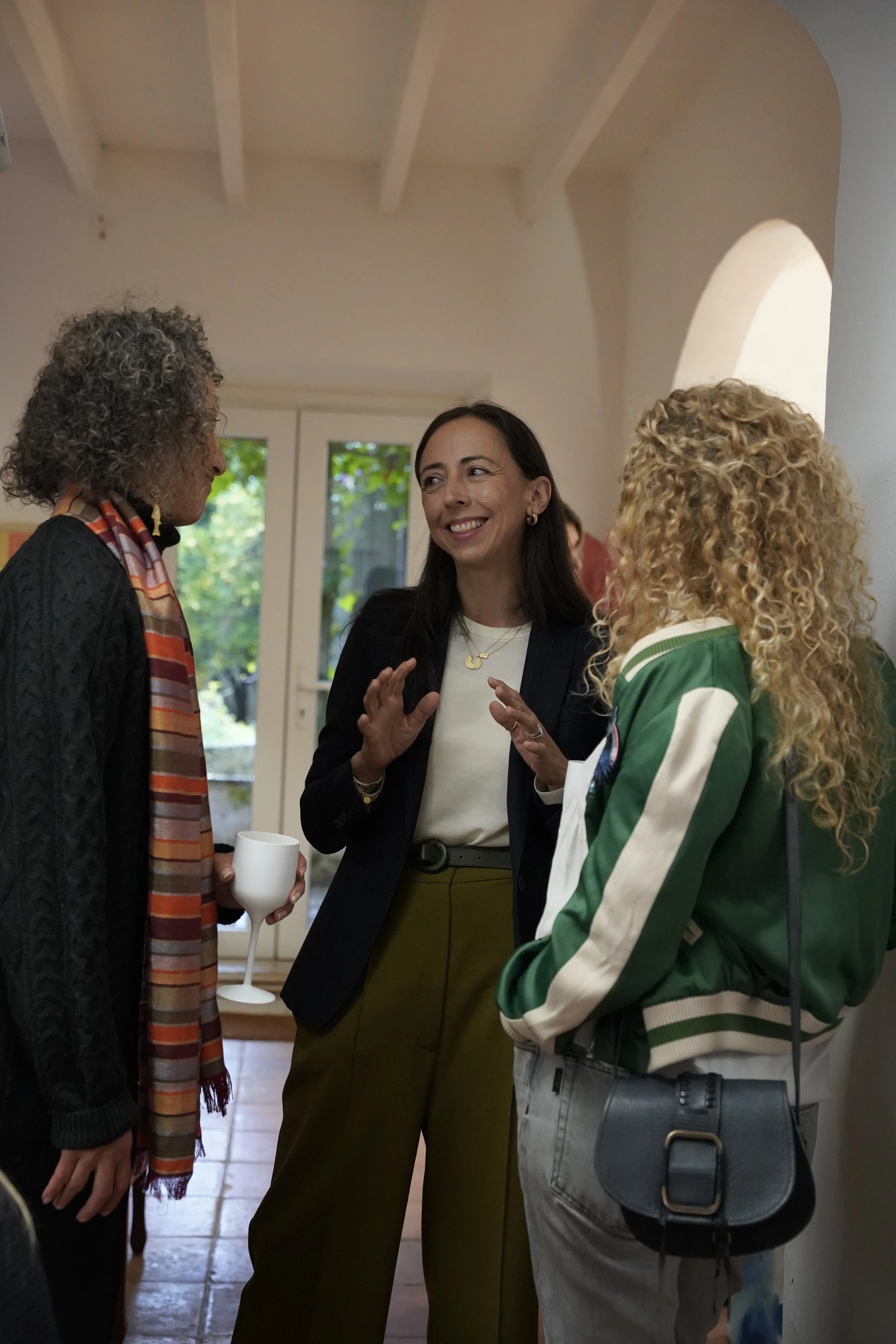 A woman with dark hair talking to two women with curly hair in a kitchen or dining area, with two windows and greenery outside.
