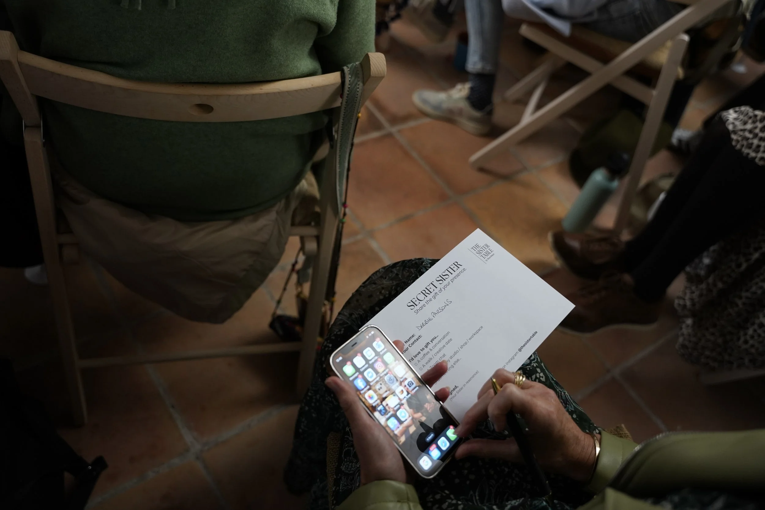 Person holding a smartphone with a colorful app grid, sitting next to a booklet titled 'Secret Sister' on their lap, in a casual indoor setting with tiled floor and chairs.