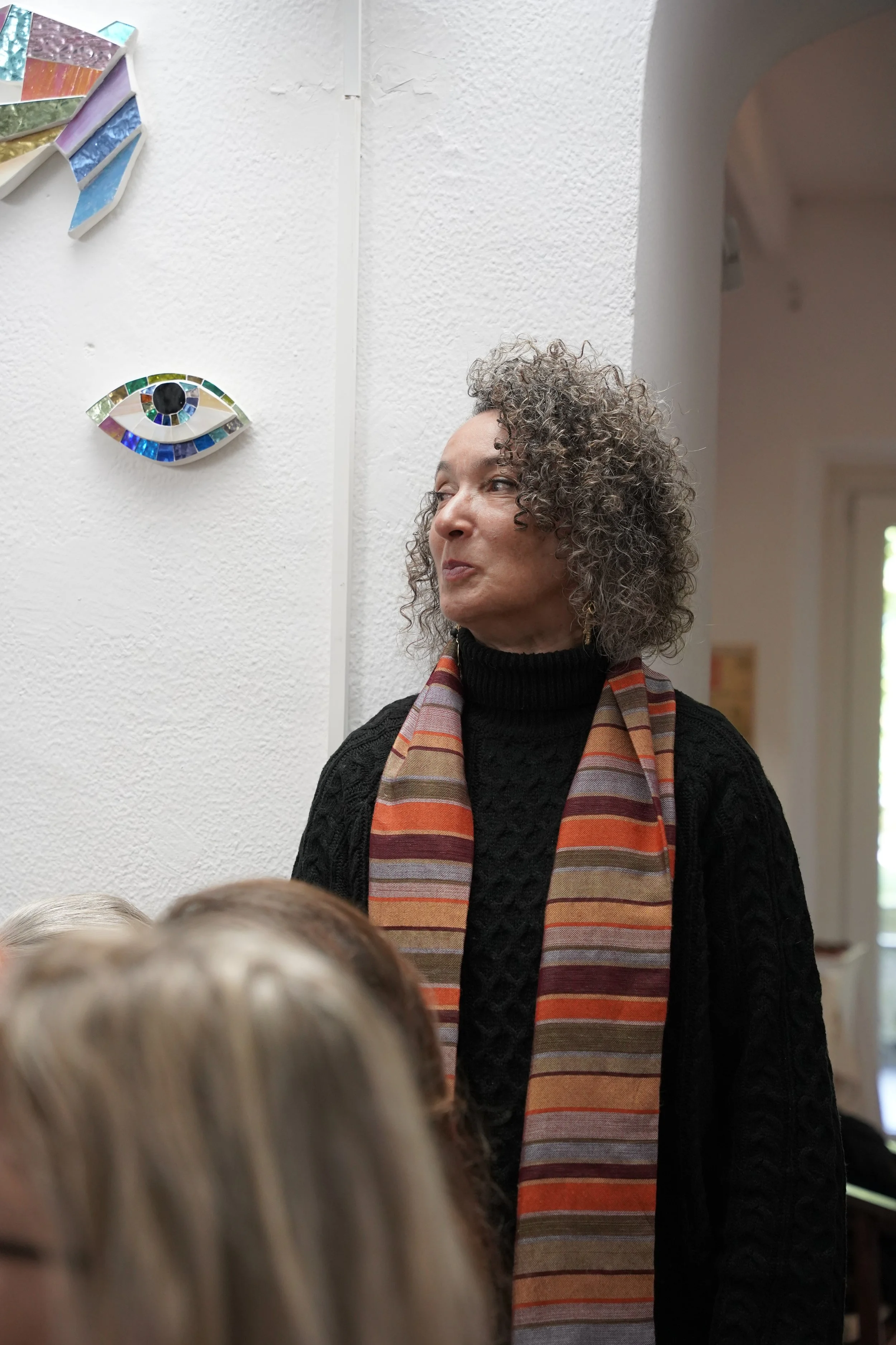 A woman with curly gray hair wearing a black sweater and a colorful striped scarf, standing near a white textured wall with mosaic art and a decorated eye-shaped mosaic above her, and a doorway visible in the background.